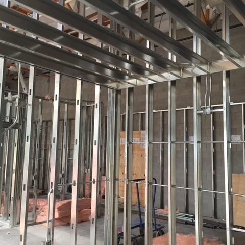 An interior construction photo focusing on the framing of a room with metal studs. The concrete block wall is visible behind the new framing. Exposed pipes and electrical conduits run through the framed walls and ceiling.