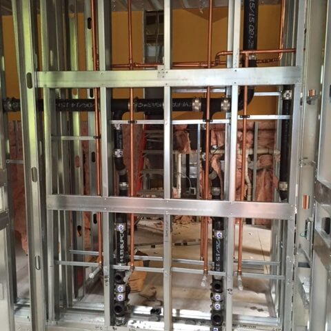 A close-up view of a framed wall section during construction, showing newly installed plumbing pipes. Copper pipes and black plastic drain pipes run vertically and horizontally within the metal stud framing.