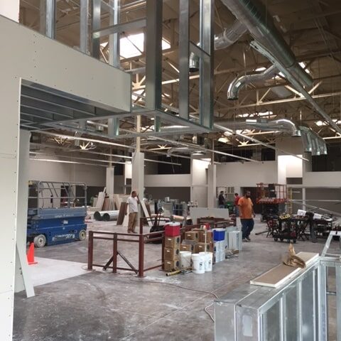 A wide shot of a large, open interior space under construction. The ceiling features exposed ductwork and light fixtures. Several workers and pieces of equipment, including a scissor lift, are scattered throughout the space. The metal stud framing for a new room is visible in the foreground.