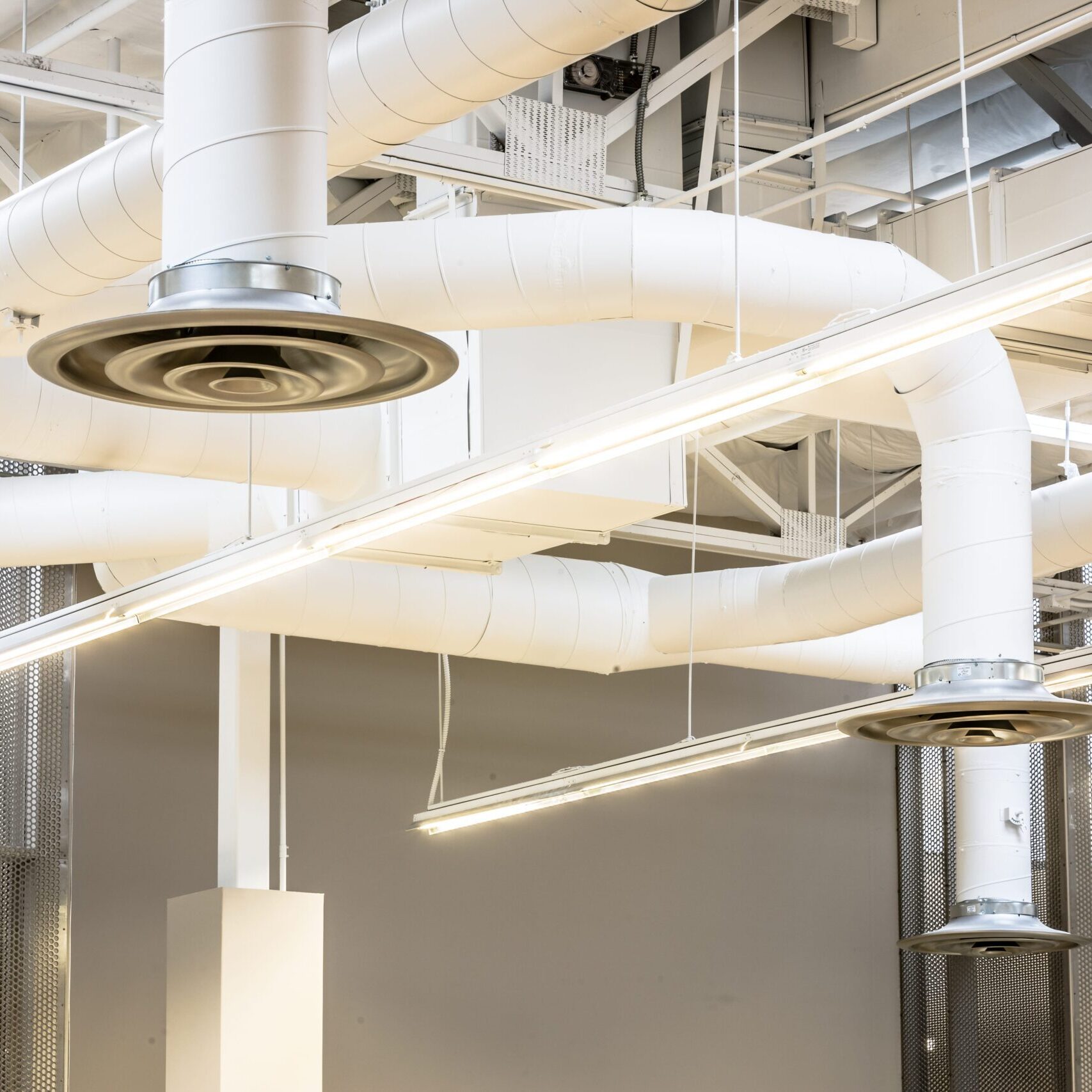 A close-up view of the white, exposed ductwork and light fixtures on the ceiling of the library. Large, winding circular ducts and vent grilles hang from the ceiling, along with long, linear LED lights.