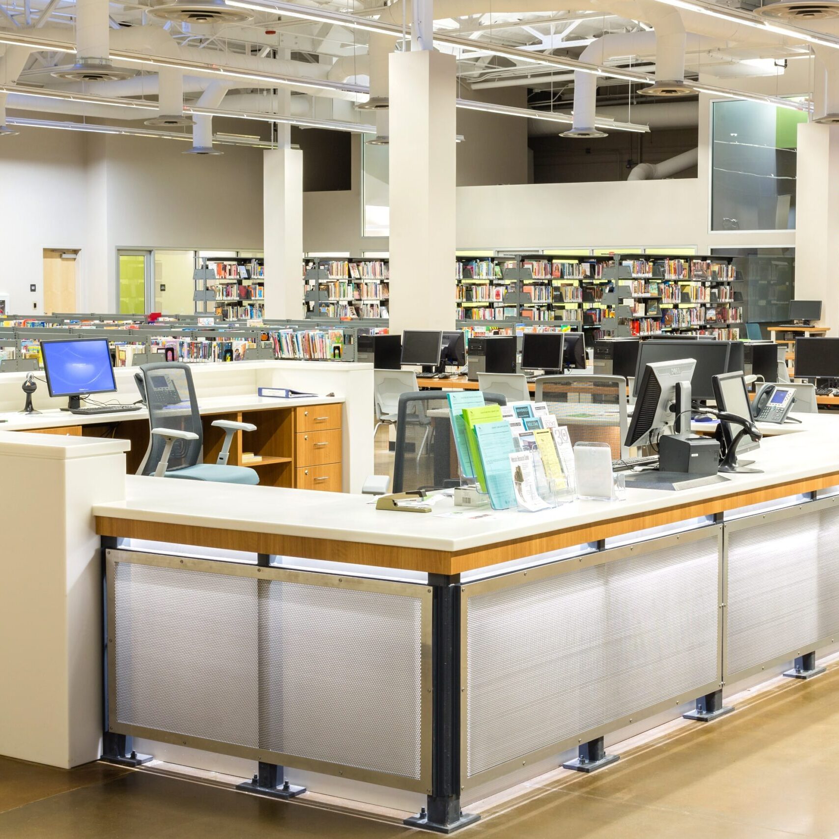 A well-lit, interior shot of a modern library's circulation or information desk area. In the foreground, there is a large, white counter with a textured, perforated metal facade and a light-colored top. A sign that reads "BOOK DROP" is visible on the left side of the counter. Beyond the counter, rows of bookshelves filled with books are visible, as well as several desktop computers on a separate table. The ceiling has exposed pipes and ductwork. On the far right wall, a large, colorful mural is displayed. The floor is a polished concrete surface.