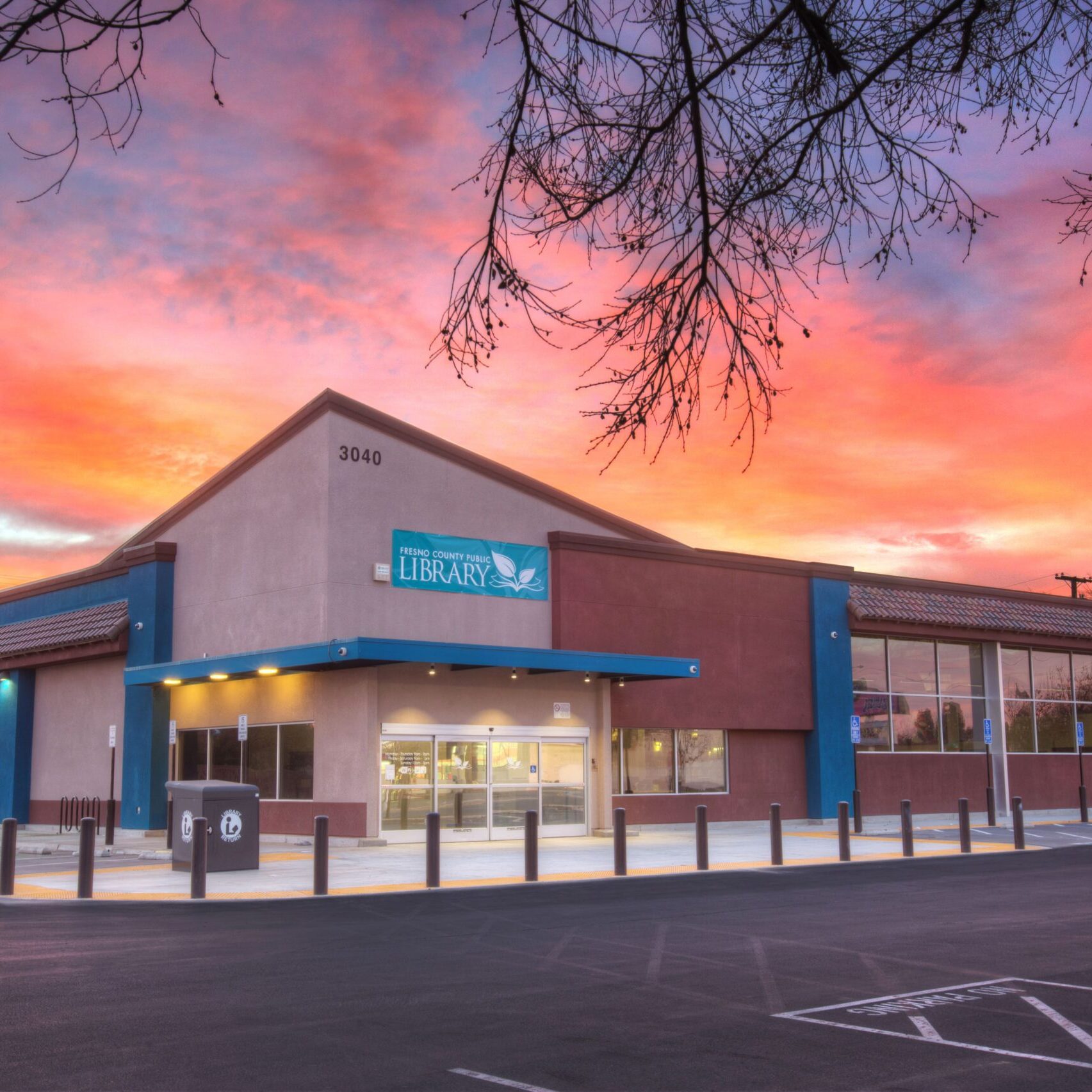 An exterior photo of the Betty Rodriguez Library at dusk. The building has a modern design with multi-toned walls and blue accents. The sky is a vibrant mix of orange, pink, and purple from the sunset, and a tree with bare branches frames the top of the photo.
