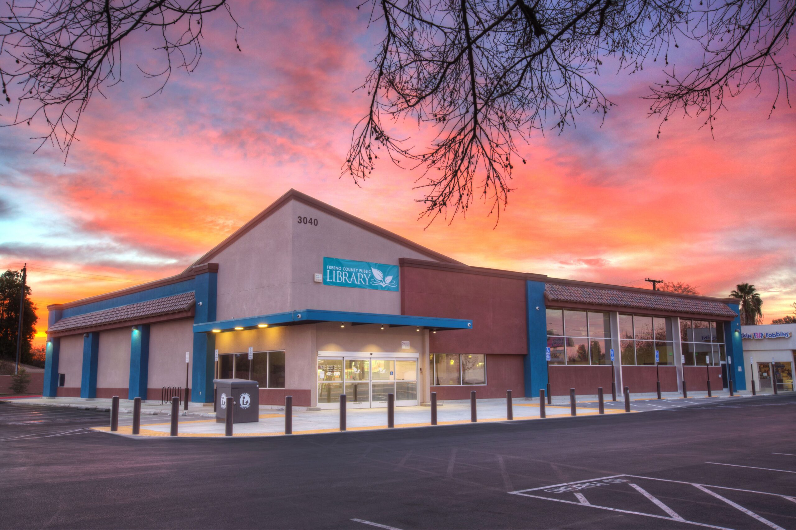 A dramatic, wide-angle exterior shot of the Betty Rodriguez Library building at sunset or sunrise. The sky is filled with vibrant hues of orange, pink, and purple clouds. The one-story library building has a contemporary design, with a light-colored central section and a darker red and blue section to the right. A blue awning with the word "LIBRARY" and an owl logo is above the main entrance. Bare tree branches are visible in the foreground, framing the top of the image. The parking lot in front of the building is empty, with black bollards lining the walkway. The building number "3040" is visible near the roofline.