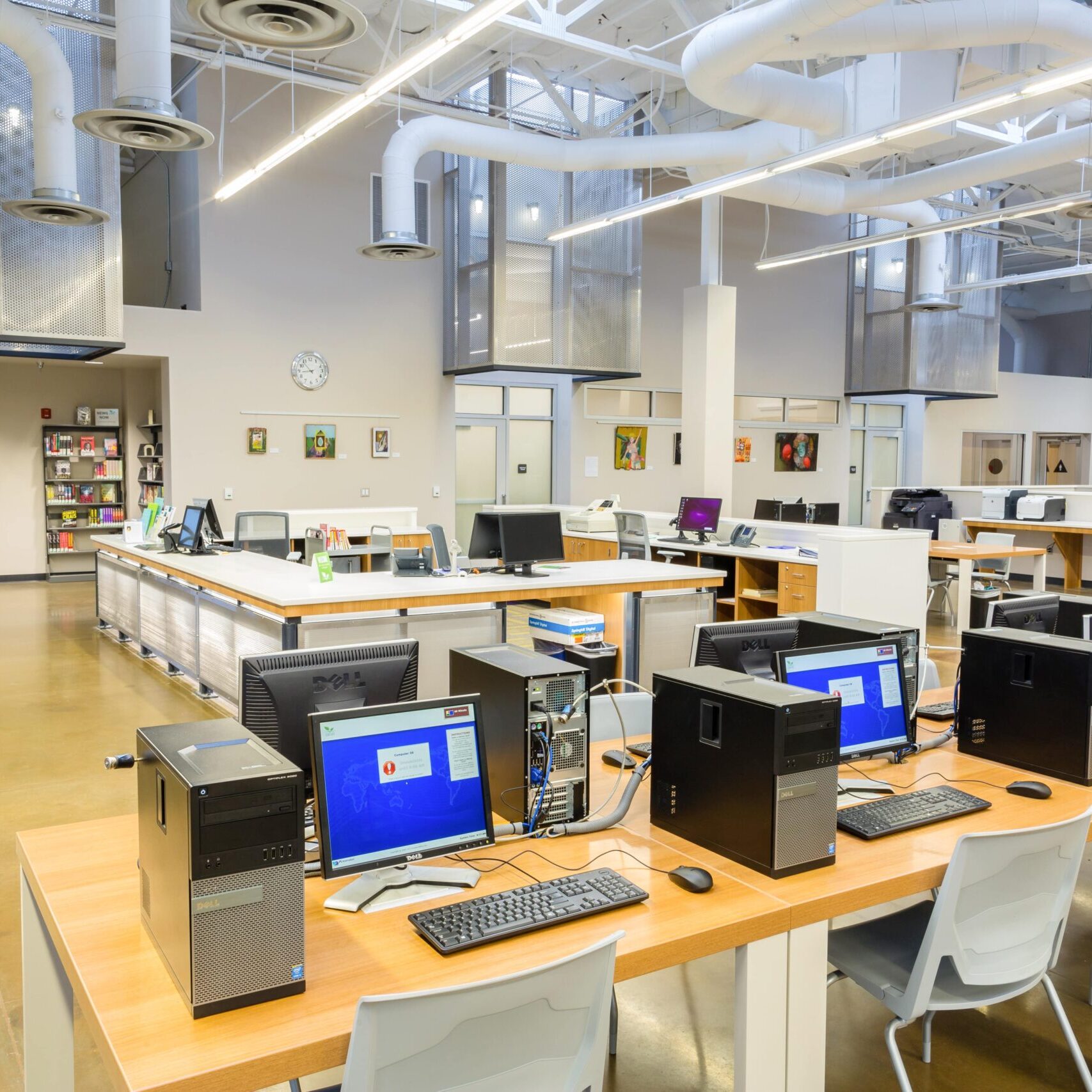 A bright, wide-angle shot of the interior of a modern public library. In the foreground, there are rows of wooden desks with multiple desktop computers, keyboards, and monitors. The computers have their monitors on, displaying a login screen. In the background, there is a large, central table, also with computers and chairs. To the left, a bookshelf is visible, and further back, there are two private rooms with glass walls. The ceiling has exposed ductwork, pipes, and fluorescent lighting. The floor is a polished concrete surface. The overall atmosphere is clean, modern, and well-lit.