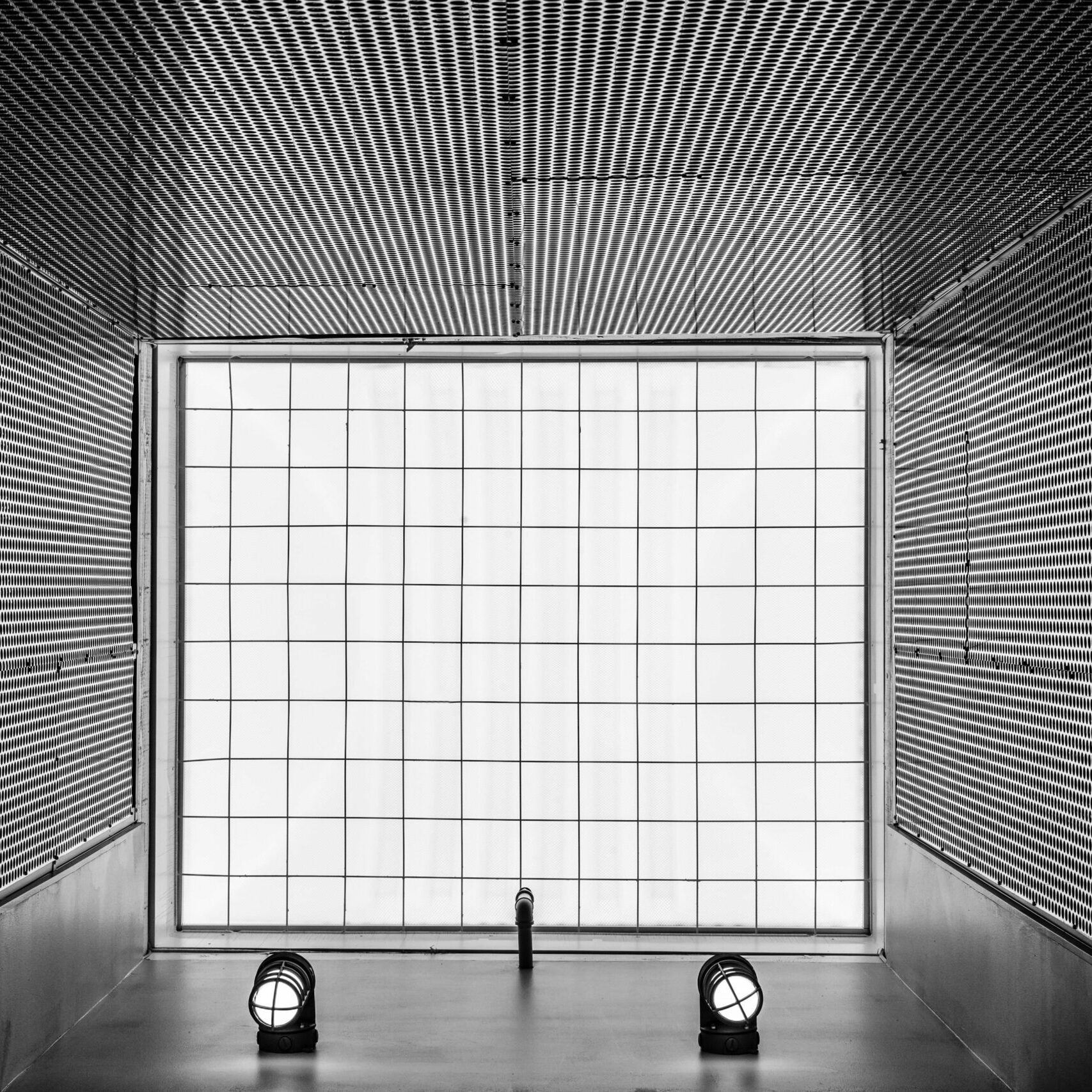 A high-contrast black and white architectural detail shot of an enclosed space. The walls and ceiling are covered with a textured, perforated metal material, and a large, gridded, frosted window panel occupies the back wall. Two cylindrical floor lights illuminate the space.