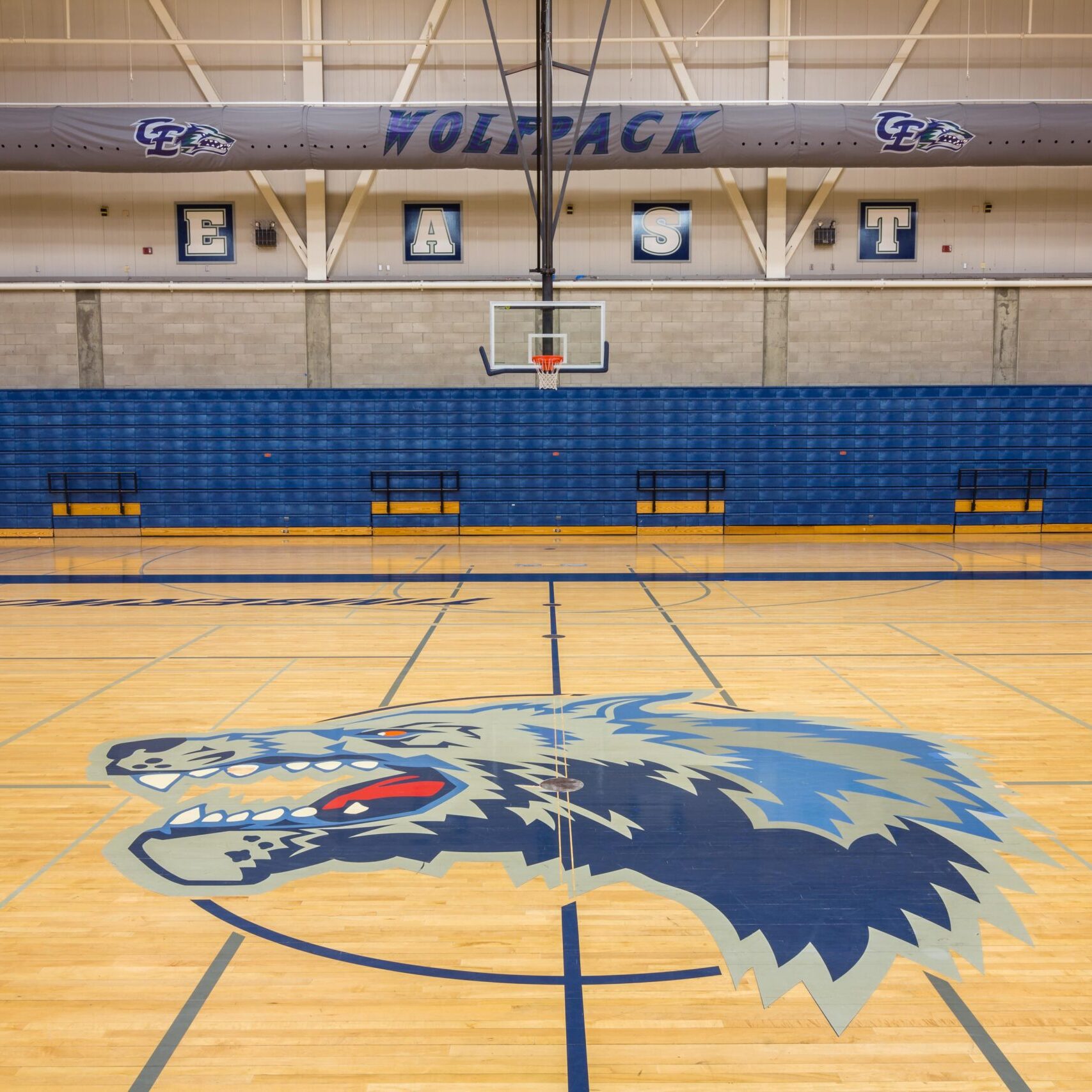 The interior of a high school gymnasium, with a large, stylized wolf head logo painted on the center of the wooden basketball court. The logo is dark blue, light blue, and gray. The bleachers on the side are retracted and blue. A large banner with the word "WOLFPACK" is hanging above the bleachers.