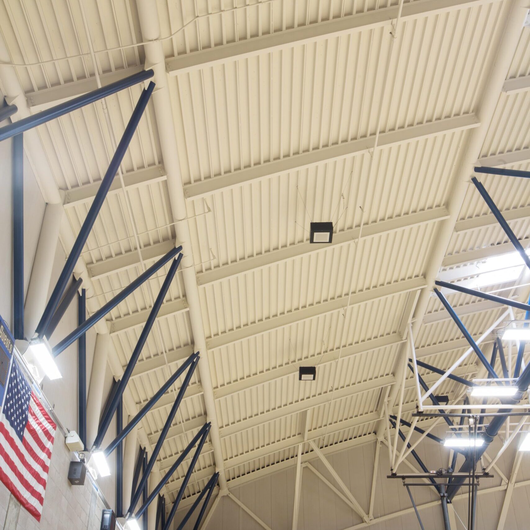 A low-angle shot showing the ceiling and upper walls of a gymnasium. The ceiling is white with corrugated metal decking. A large American flag hangs on the wall, and parts of the blue and white steel truss system are visible.