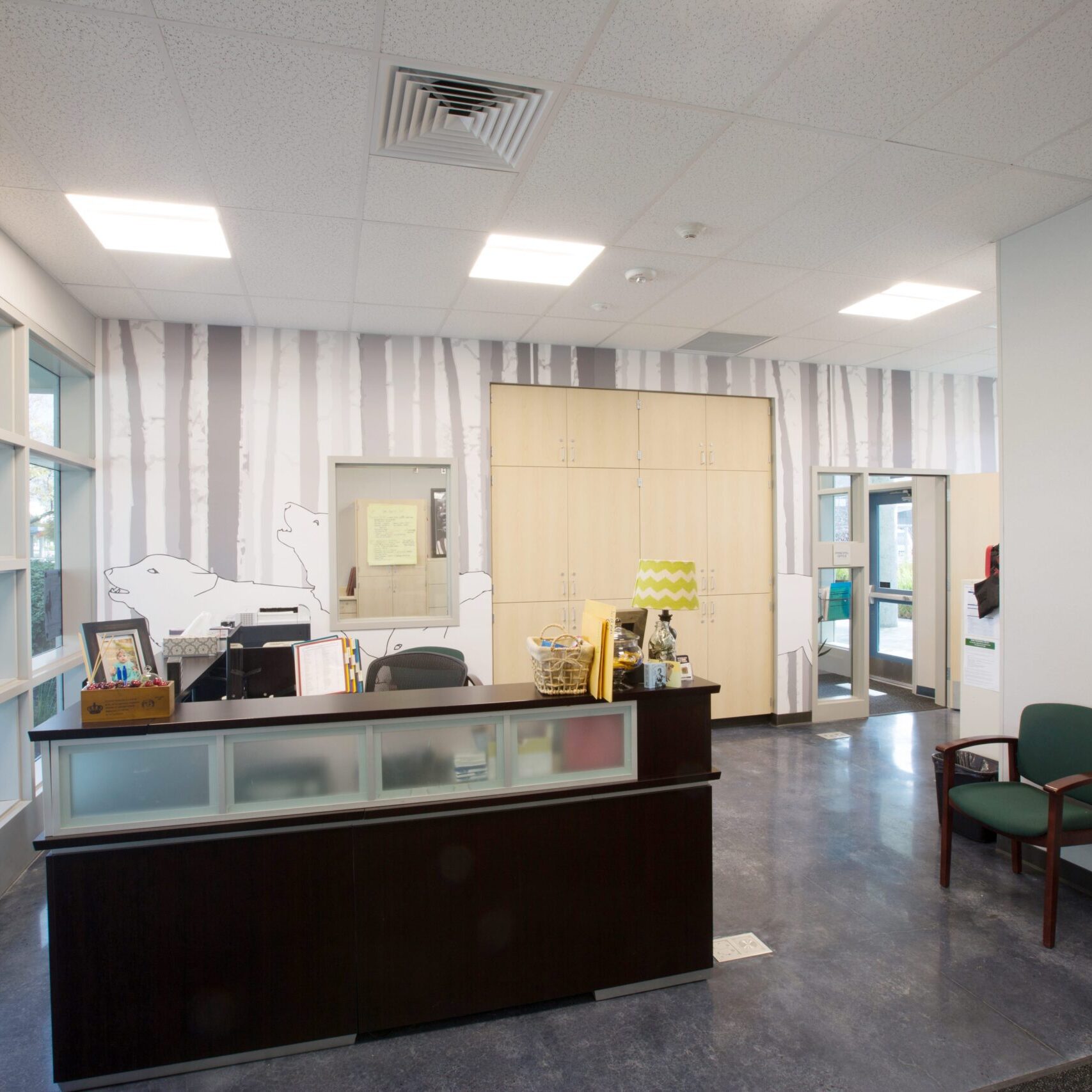 The interior of a modern school office, likely a main or front office. A dark wood and frosted glass reception desk is in the center, with a computer monitor visible. A wall with a light gray and white graphic of trees is in the background, along with a green armchair.