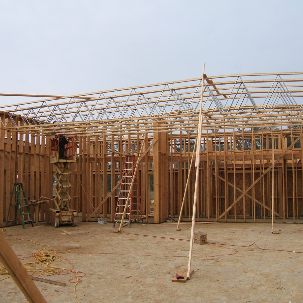 A view of the interior of a building under construction. The walls and ceiling are framed with wood and metal trusses. A worker on a scissor lift is working near the ceiling.