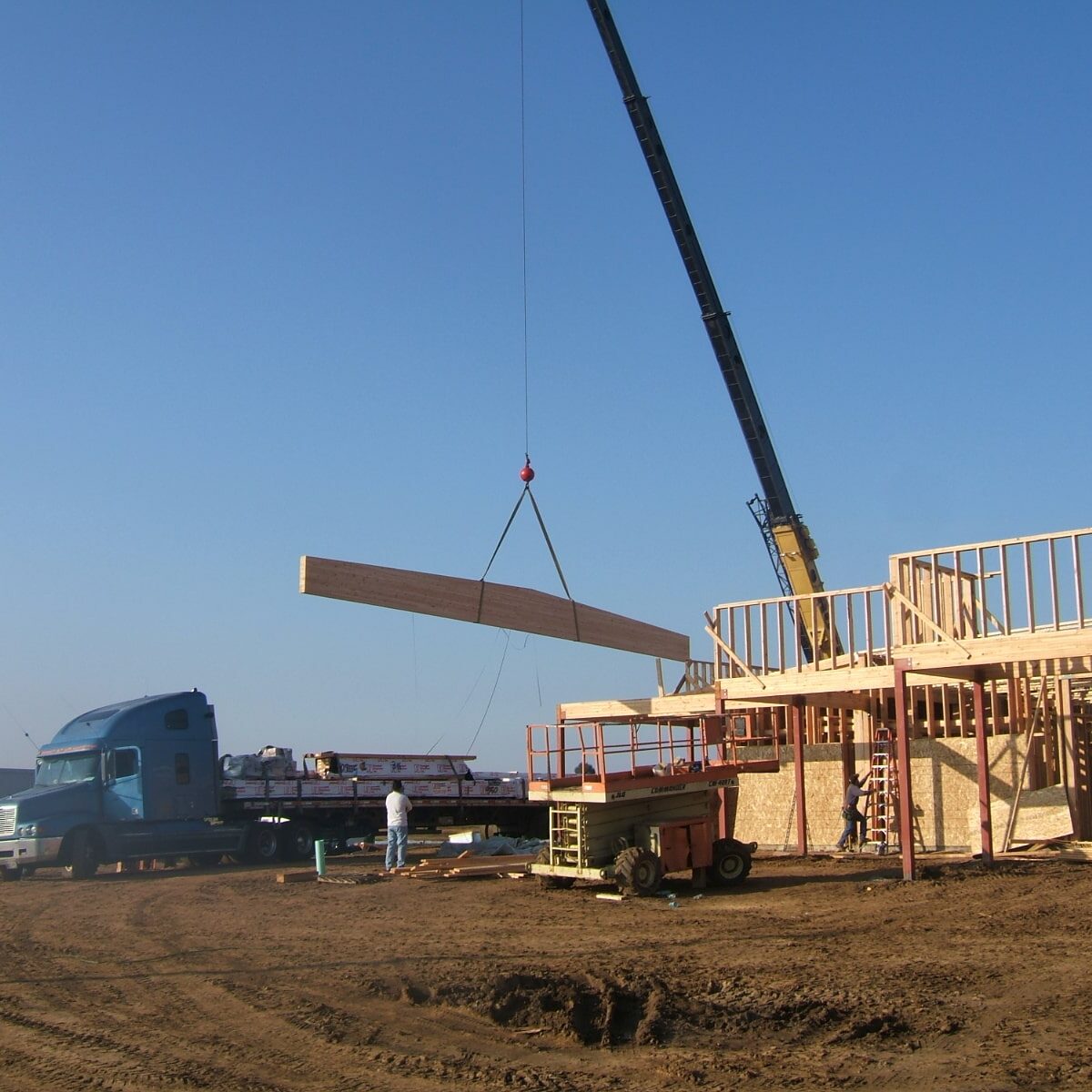 A construction site photo. A large blue truck is parked next to the framed walls of a building. A crane is lifting a long wooden beam into place. A worker stands below, guiding the beam.