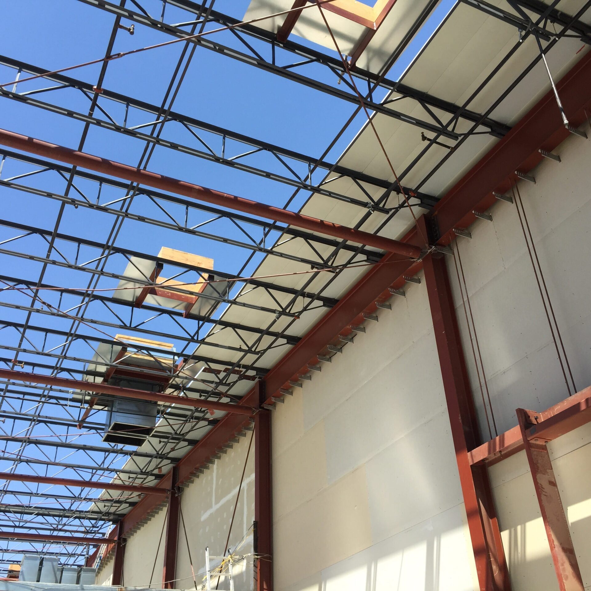 A low-angle interior shot of a building under construction, looking up at the ceiling. The ceiling is made of exposed steel beams and trusses with some panels of white material. The walls are partially covered with white siding.