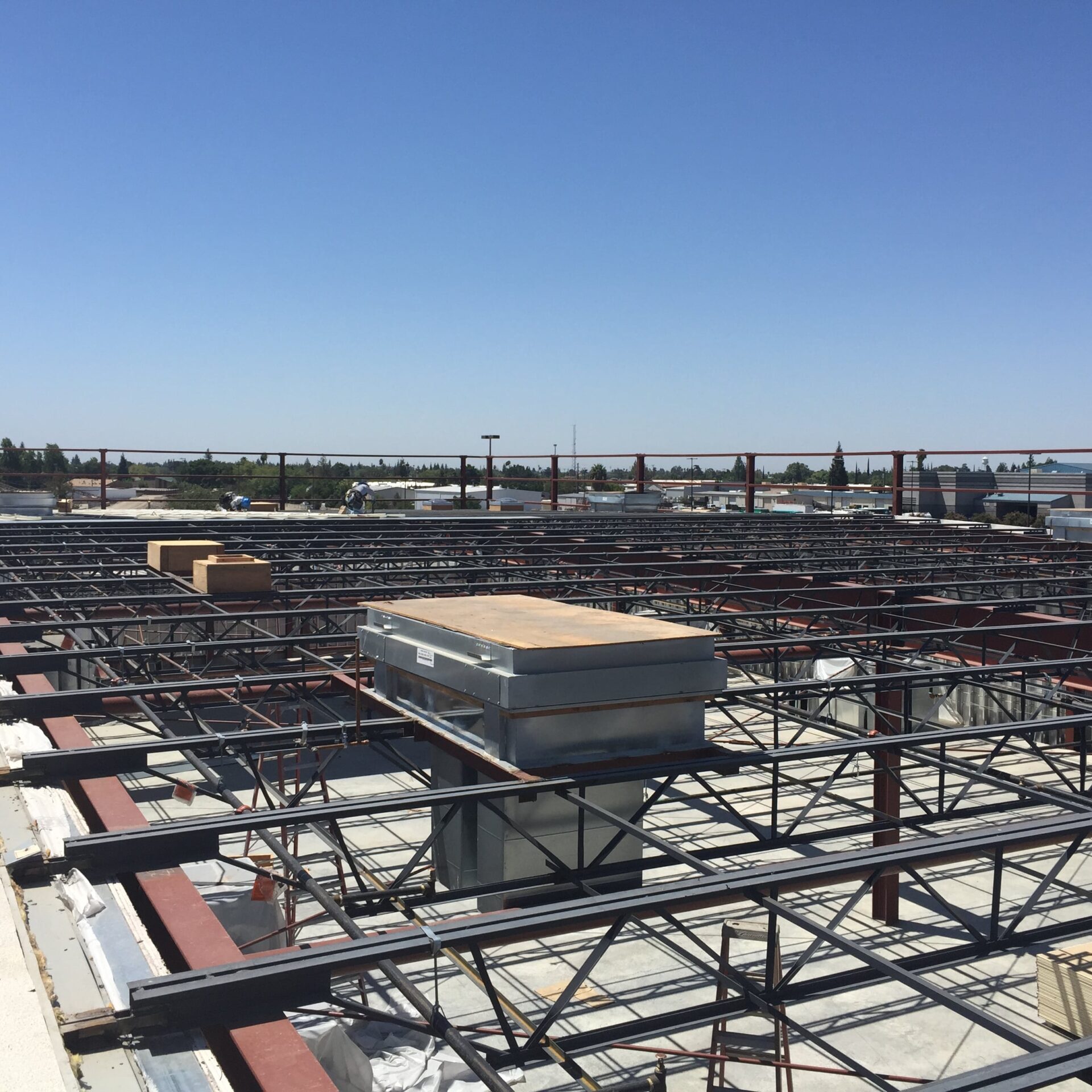 A view from the roof of a building under construction. The roof structure is made of a grid of steel joists and beams, and a large metal box-like structure sits in the middle. The horizon is visible in the background with other buildings and a clear blue sky.