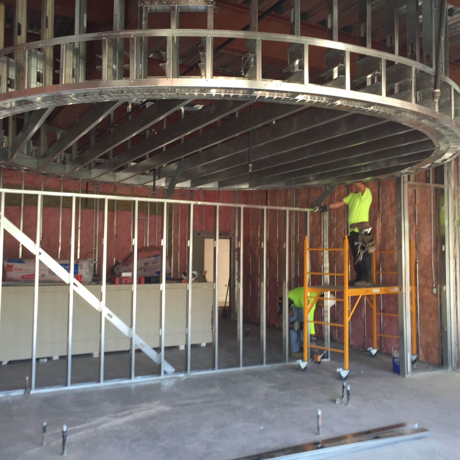 Two construction workers are inside a building, framing an interior space. One worker stands on a small scaffold, installing a curved metal frame in the ceiling. The walls are framed with metal studs and pink insulation.