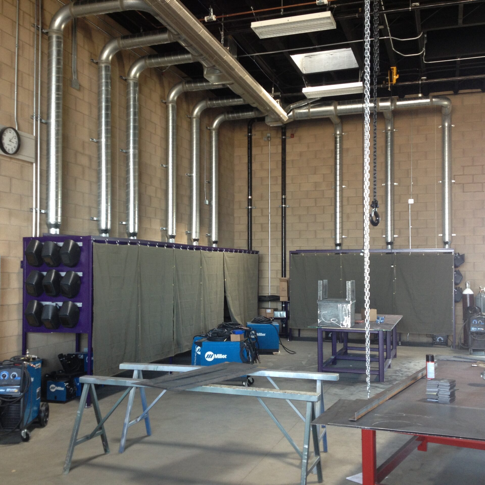A wide shot of a welding workshop. The walls are made of light brown brick and the floor is concrete. On the left are several welding machines, and on the right is a large metal workbench with various metal pieces on it. A chain hangs from the ceiling in the center. In the background, there are two covered booths with welding machines and a large system of metal pipes and ducts on the wall.
