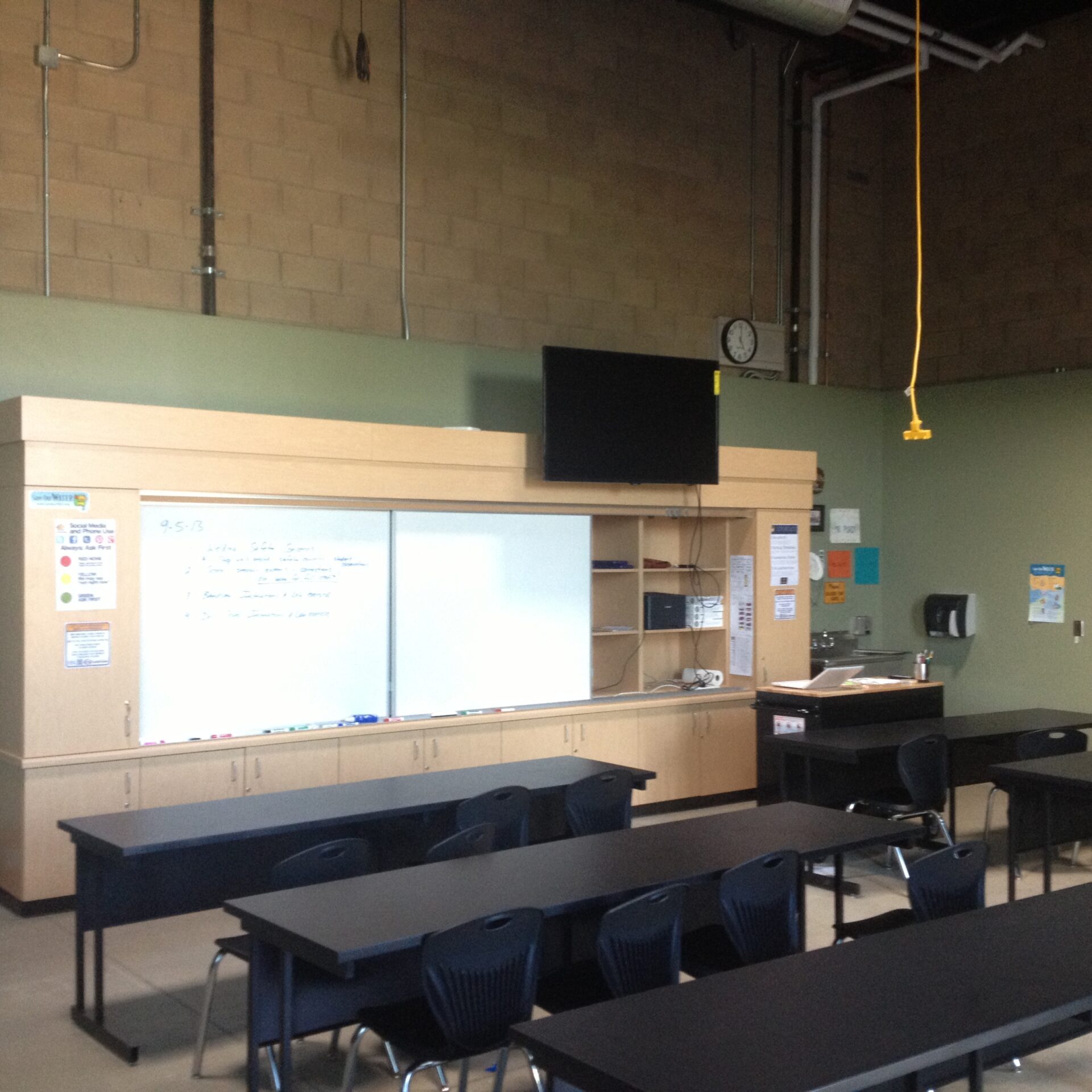 A classroom with rows of black desks and chairs facing a large whiteboard. The walls are painted two tones, with a light green color on the bottom and light brown brick on top. Above the whiteboard is a large, flat-screen TV mounted on a wooden cabinet system that runs the length of the wall.