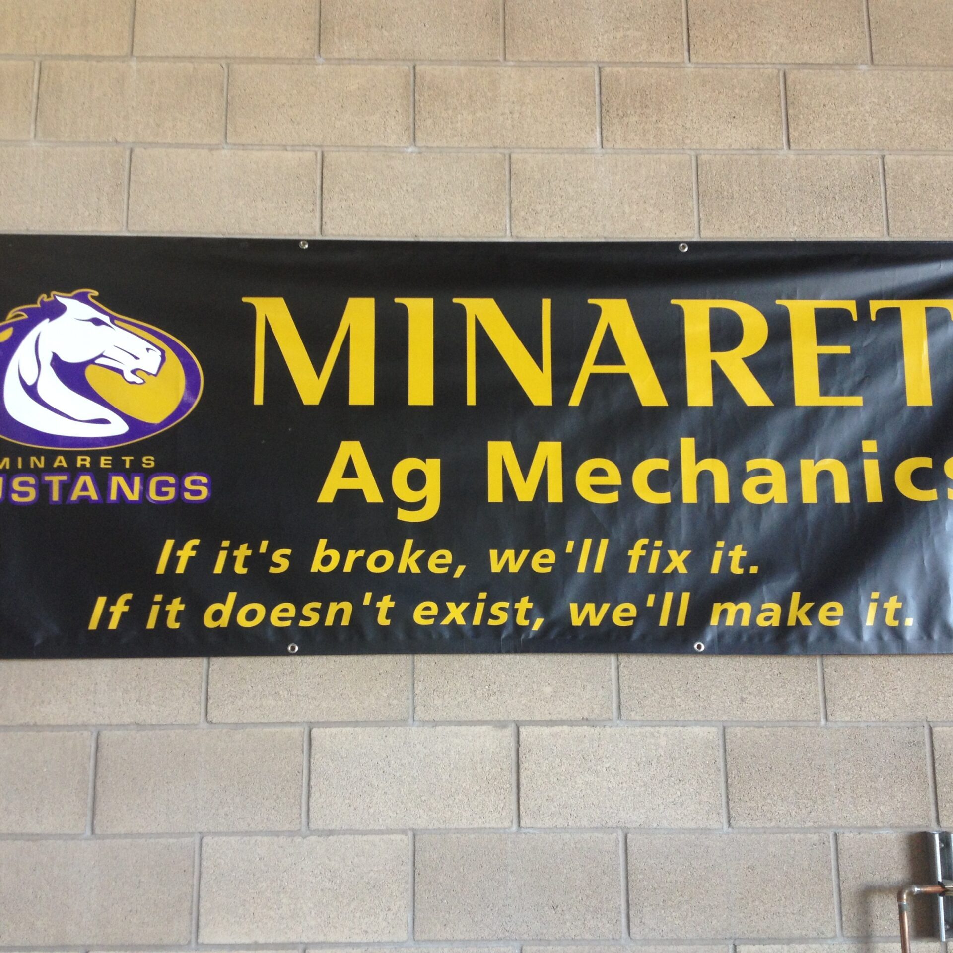 A black banner on a brick wall with the words "MINARETS Ag Mechanics" in yellow letters. To the left is a white logo of a horse with "MINARETS MUSTANGS" underneath. Below the main text are two yellow lines: "If it's broke, we'll fix it. If it doesn't exist, we'll make it."