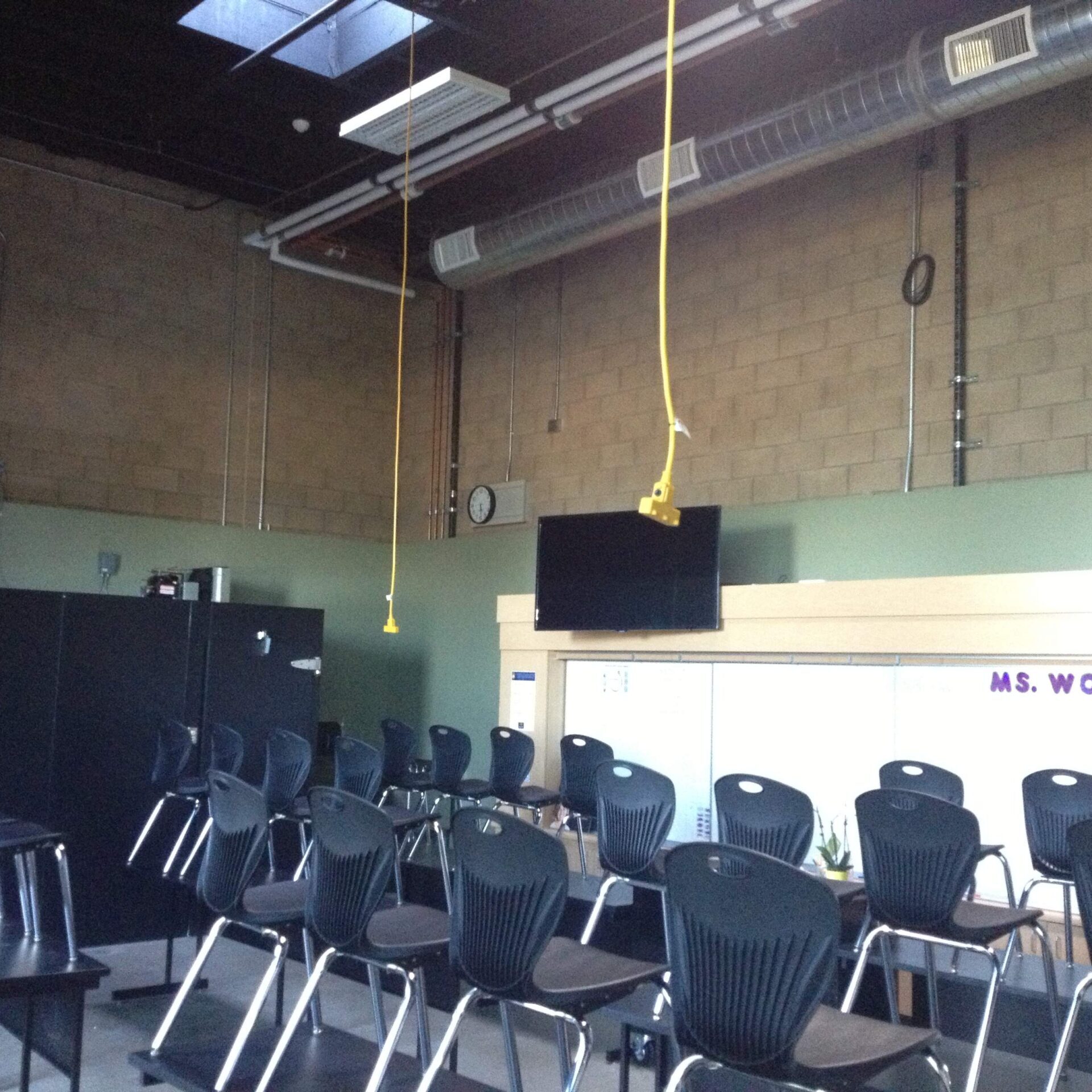 A slightly angled, wide-angle interior shot of a modern classroom or meeting space. In the foreground, there are rows of black chairs with slatted backs, facing a large white board. A television monitor is mounted above the whiteboard, which has the name "MS. WOOD" written on it. The walls are a mix of light green and unpainted cinder blocks. The ceiling has exposed ductwork and light fixtures, with several yellow ropes hanging down. The room is mostly empty of people, and several black storage cabinets are visible on the left side of the room.