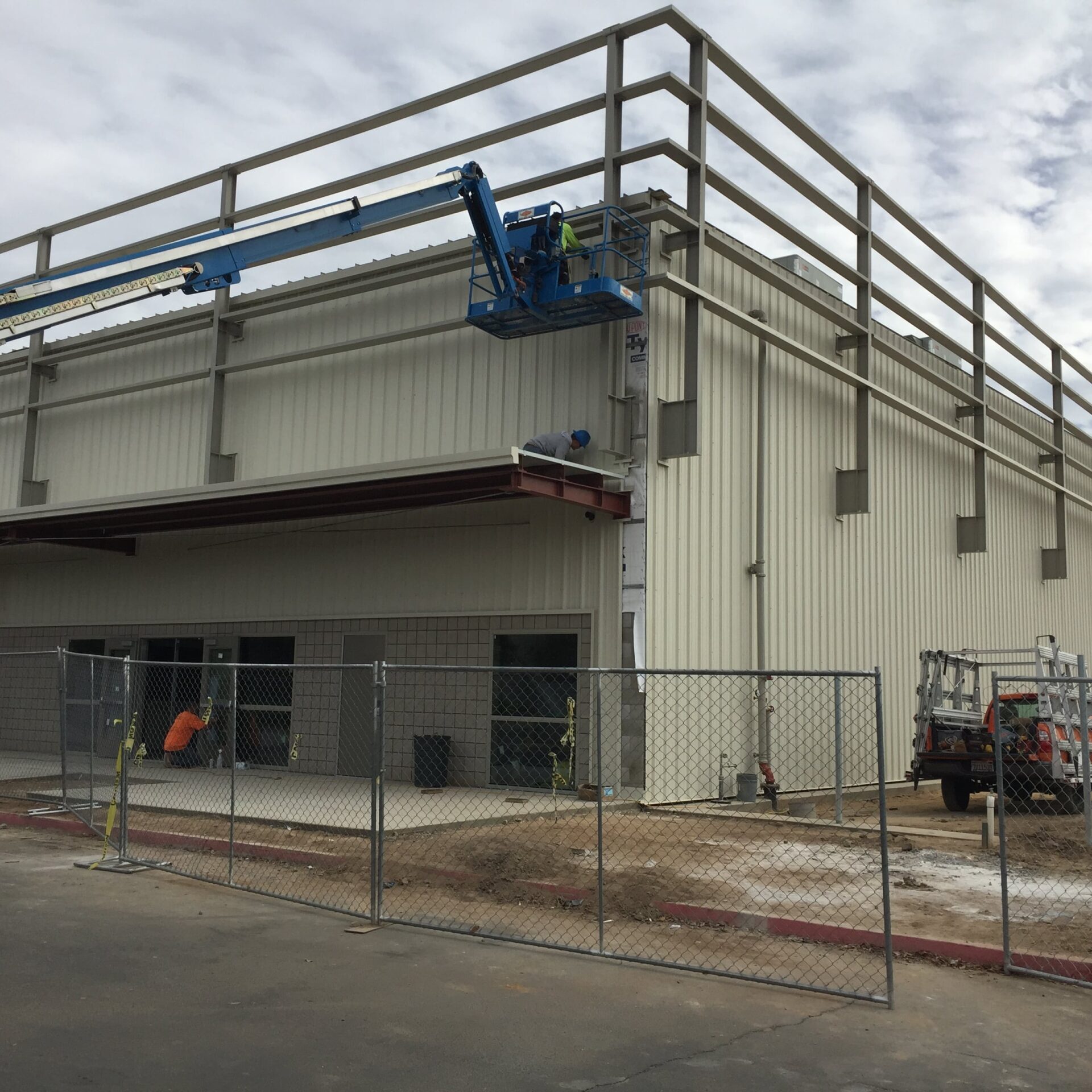 The exterior of a large, light-colored building is under construction. Workers on an aerial lift are installing a balcony or awning. A chain-link fence surrounds the building, and a pickup truck is parked nearby.