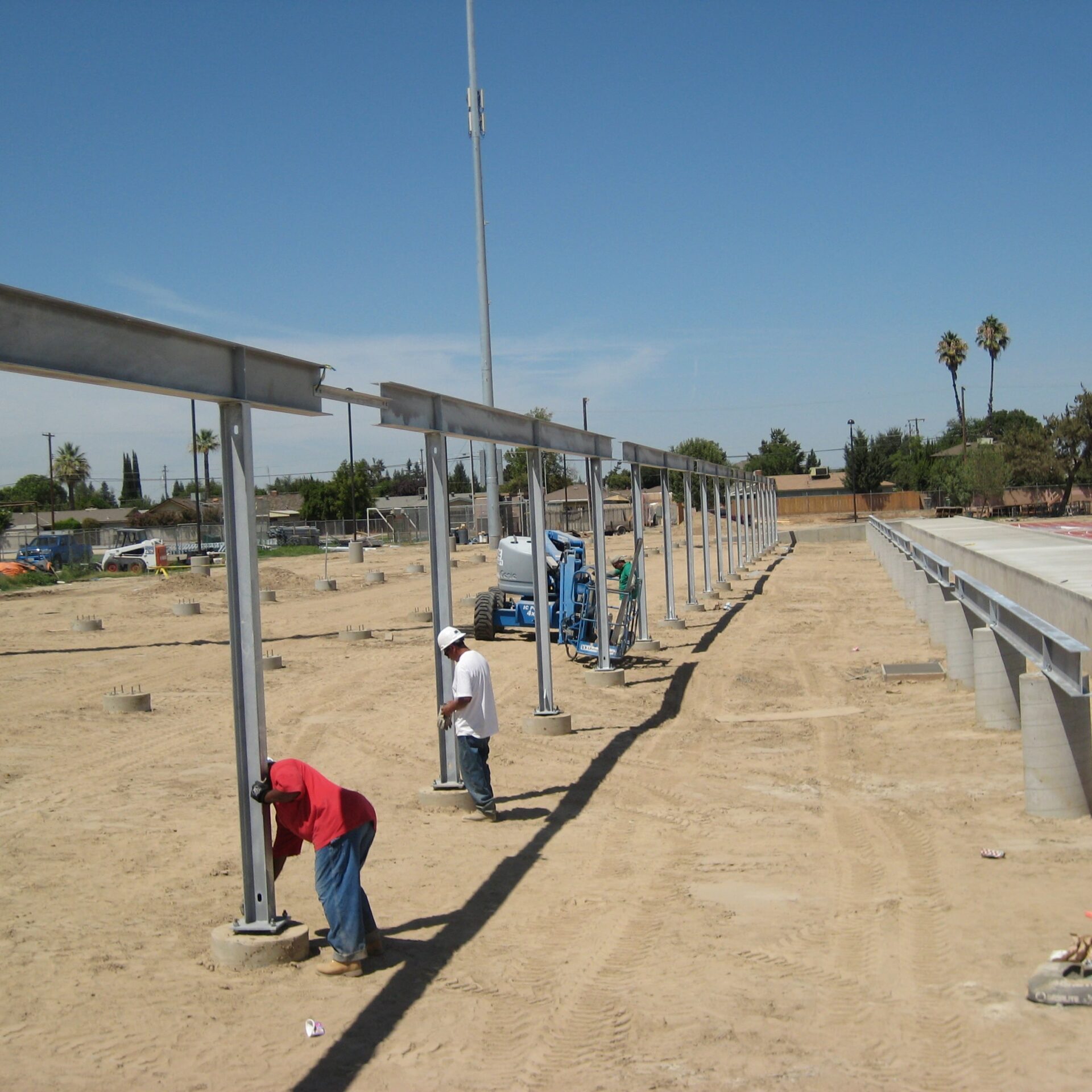 A construction crew installs the steel framework for the new grandstands at Kingsburg High School's football stadium. The framework consists of large steel beams resting on concrete foundations. The ground is dirt, and a lift machine is visible in the background.