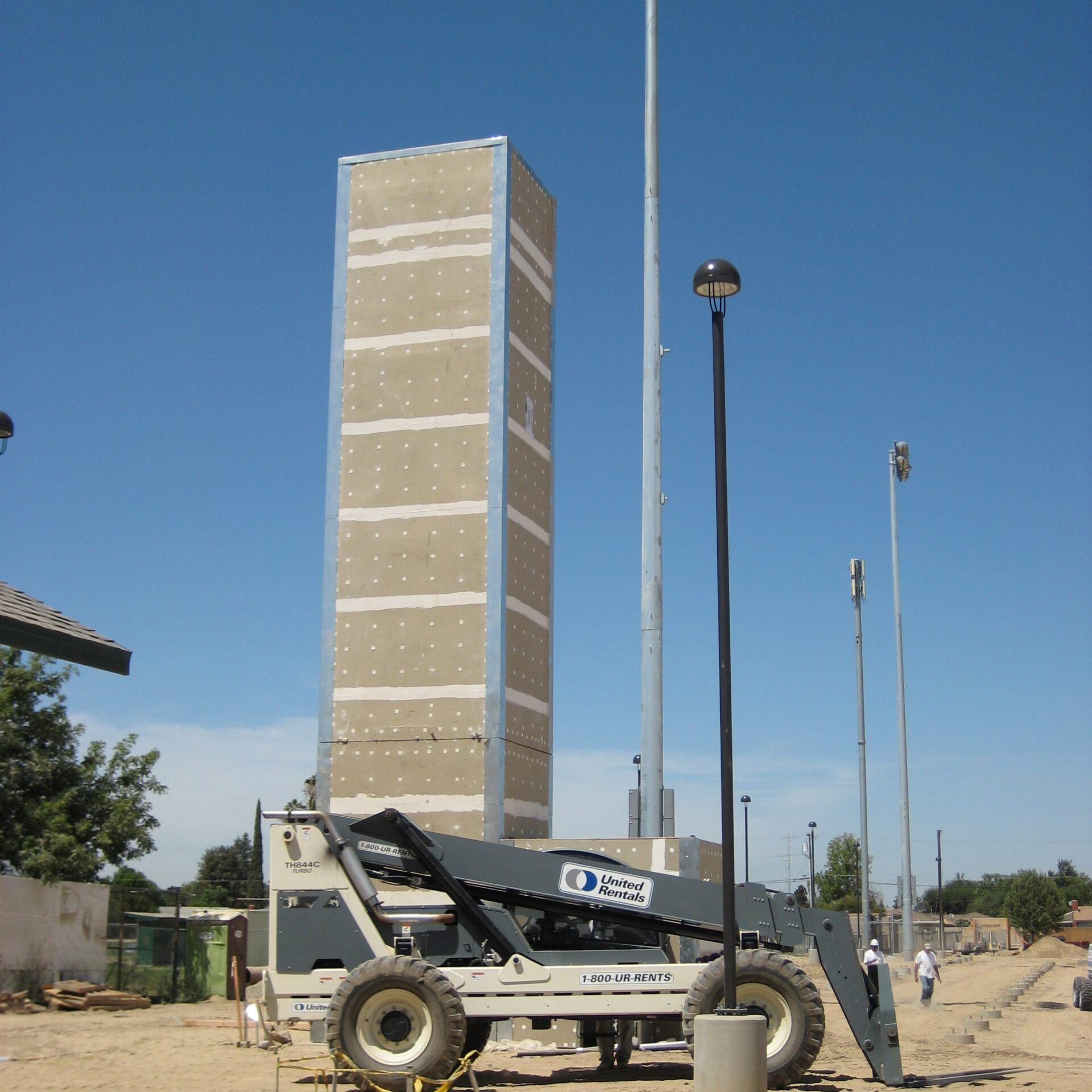 A large, rectangular building covered in what appears to be drywall or siding stands next to a tall light pole with stadium lights. A large "United Rentals" lift machine is parked in front of the building. The ground is dirt and there are other light poles in the background.