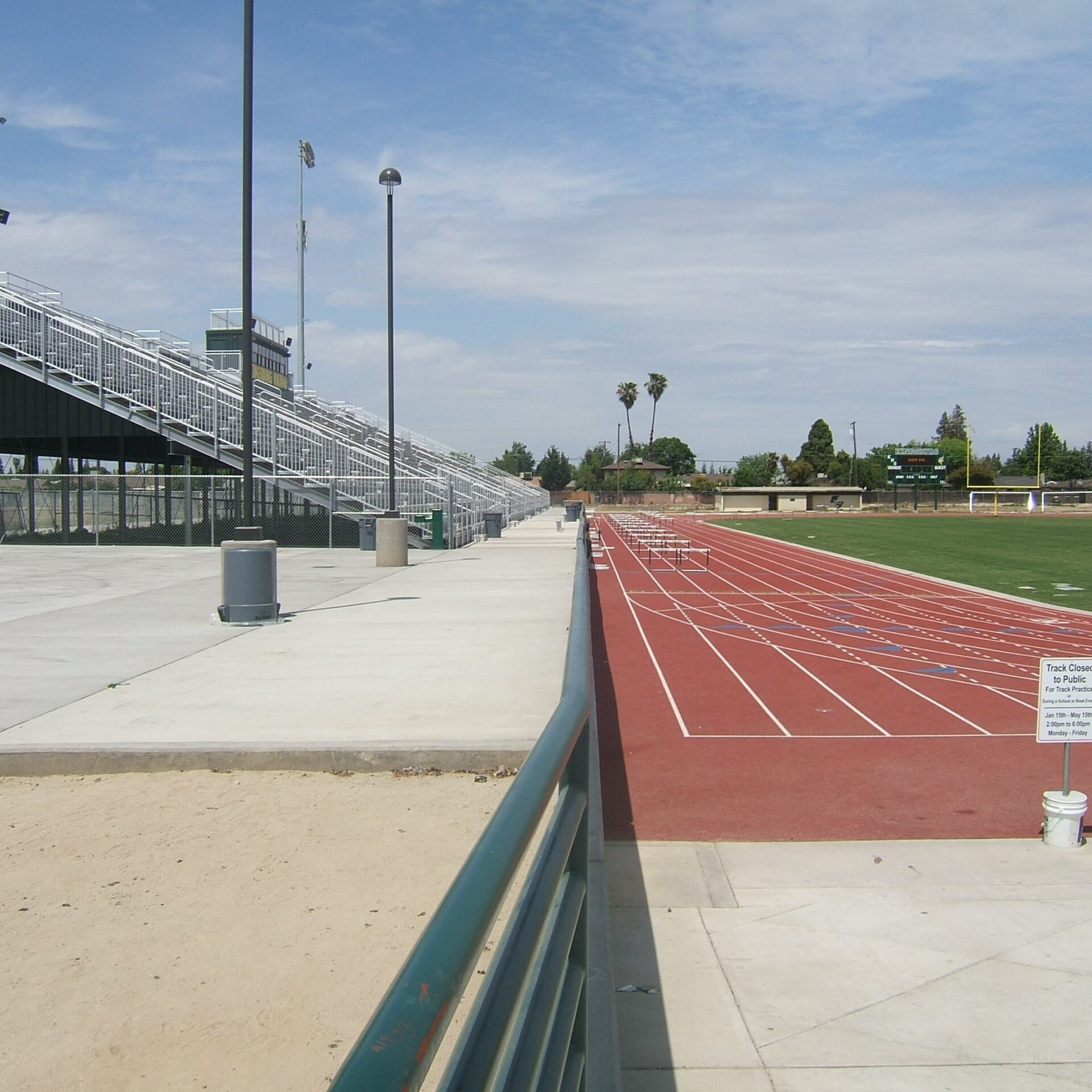The newly completed track and grandstands at Kingsburg High School's football stadium. The red running track is marked with white lines and lane numbers. A large, grey metal grandstand with multiple rows of bleachers is visible on the left. The field is green grass. A small sign on a bucket says "Track Closed".