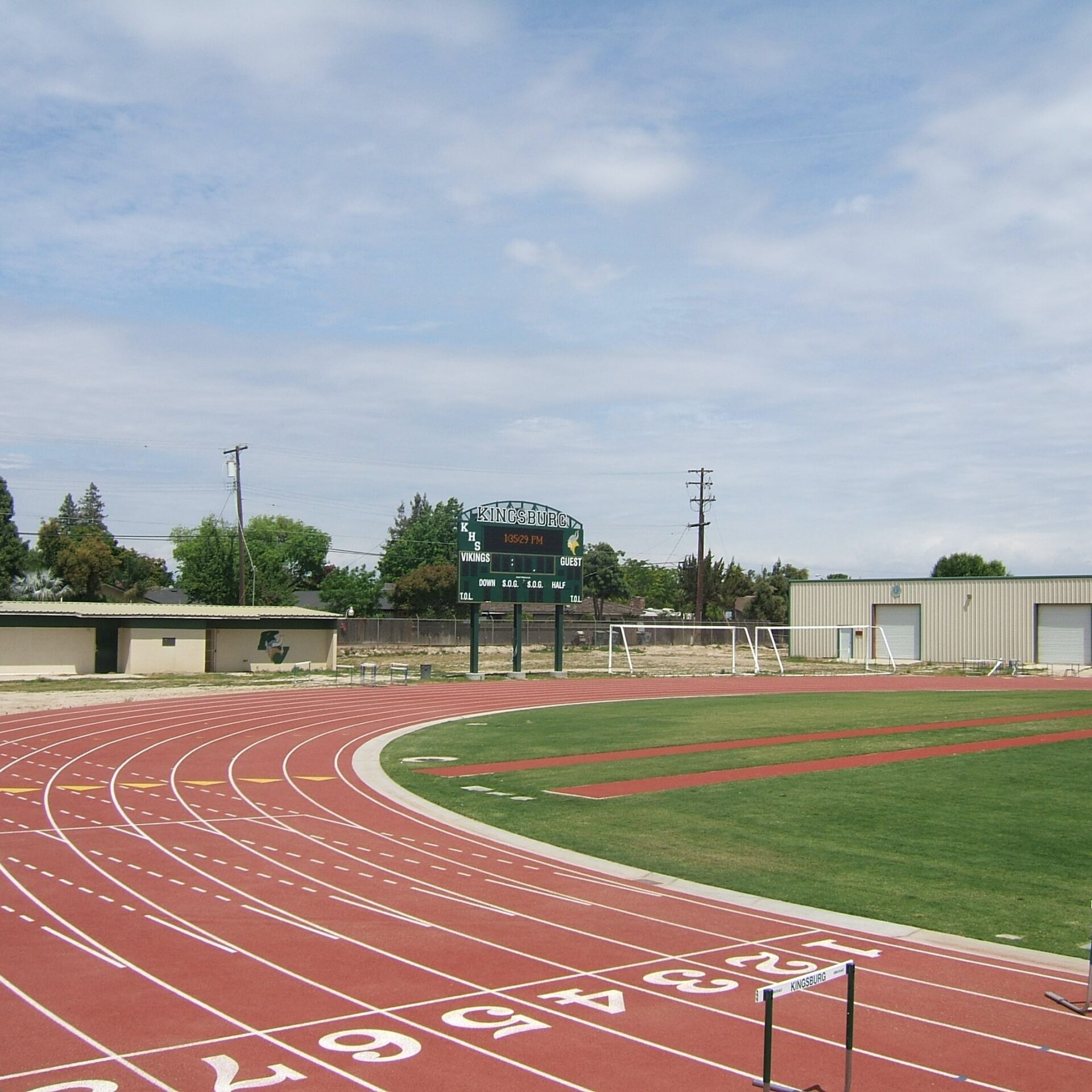 The curved portion of the new red running track at Kingsburg High School's football stadium. The track lanes are marked and there is a hurdle on the track. In the background, there is a scoreboard with "VIKINGS" and "GUEST" visible, along with a building and goalposts.