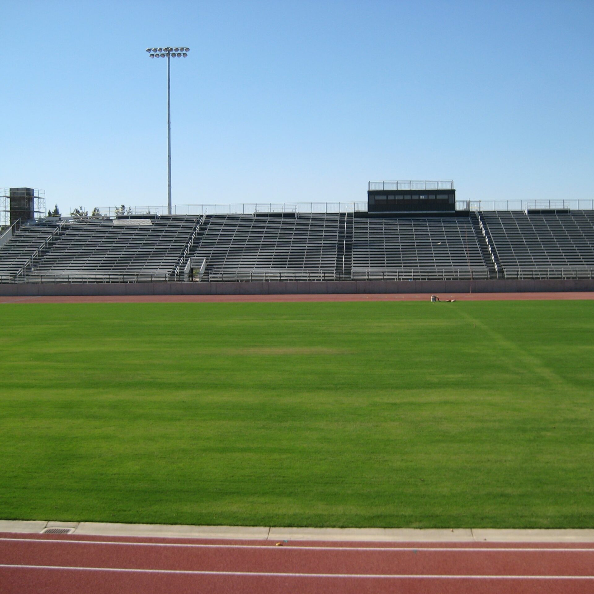 A view of the newly completed football field and grandstands at Kingsburg High School's football stadium. The green turf of the football field is in the foreground, with the large grandstands and the press box in the background. Tall light poles with stadium lights are visible on either side.