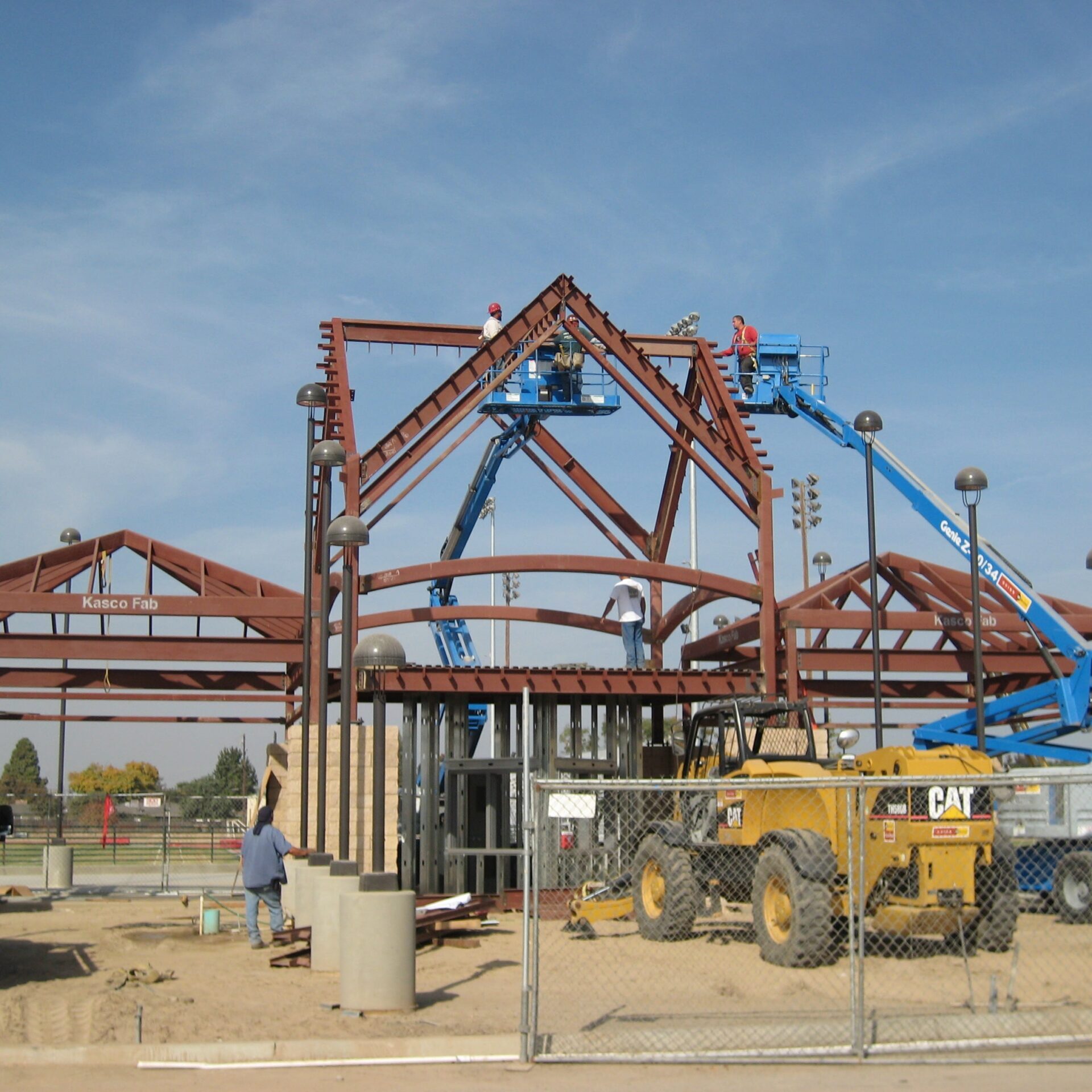 Construction workers on two aerial lifts are installing the red steel framework for a new building at Kingsburg High School's football stadium. The structure has a prominent gabled roofline. A "Caterpillar" grader is parked in front of the building. The sign "Kasco Fab" is visible on a beam.