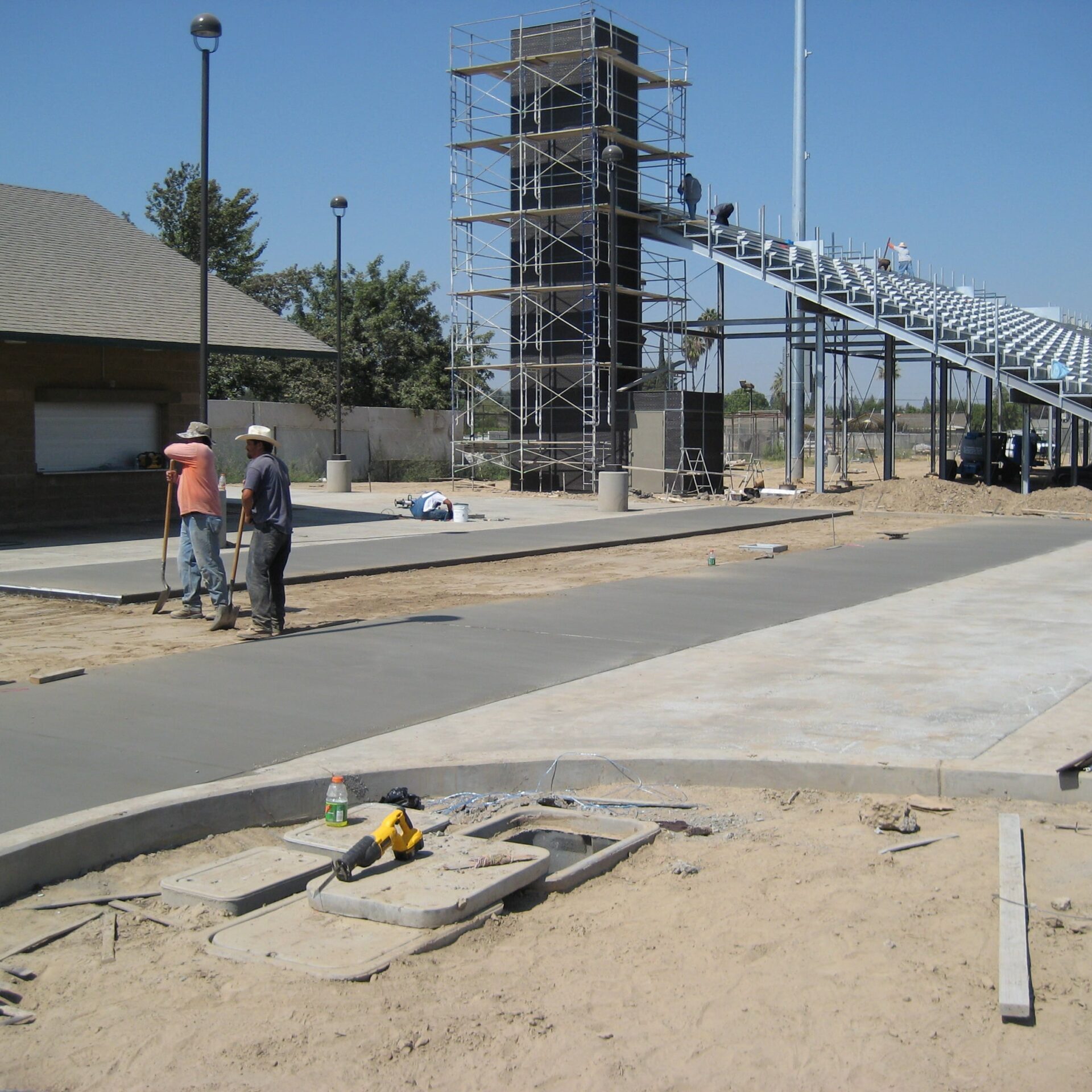 A construction scene at Kingsburg High School's new football stadium. Two workers are standing on newly poured concrete. In the background, there is a large building with scaffolding on it, a portion of the grandstands, and a smaller building with a gabled roof.