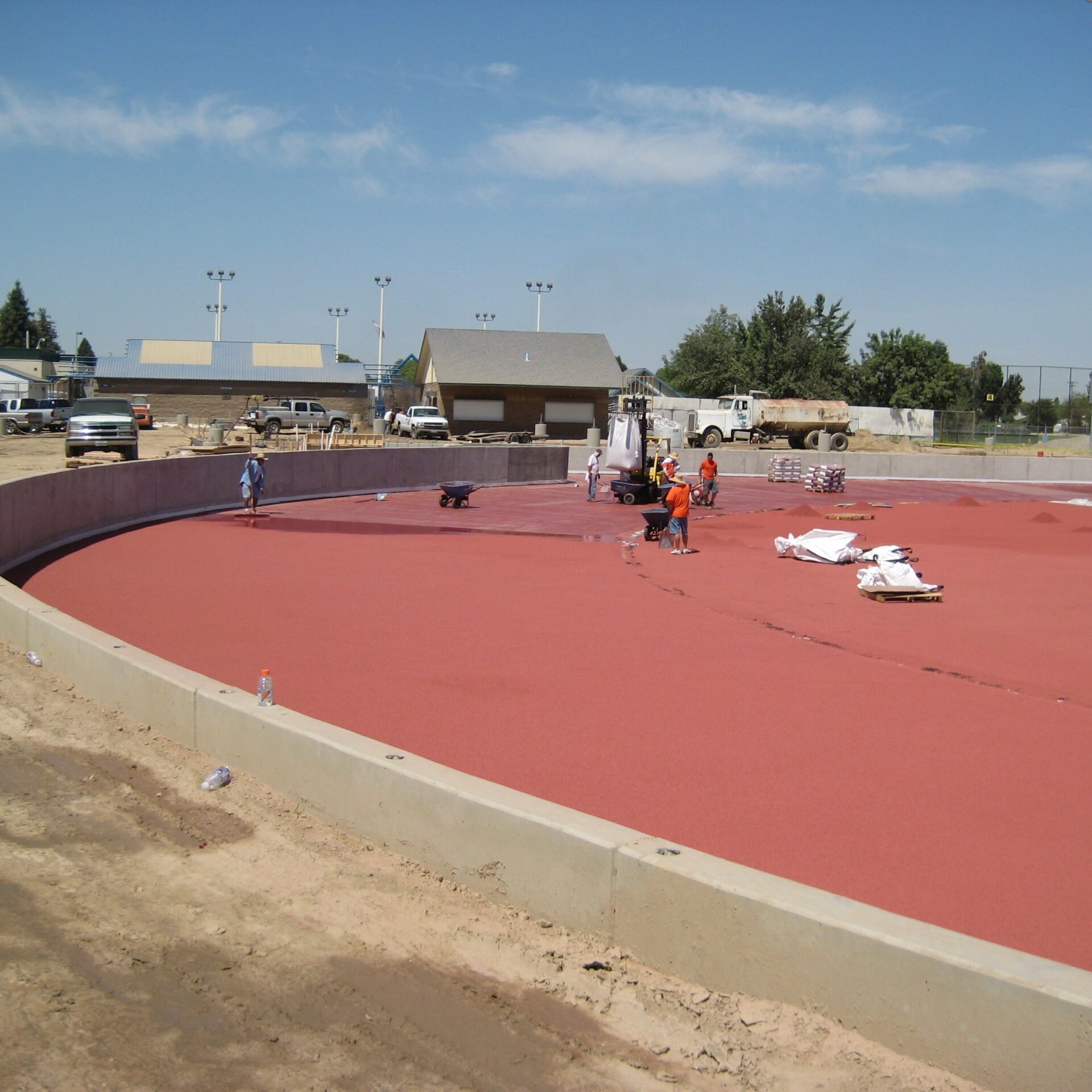 A wide view of the new red running track at Kingsburg High School's stadium during its installation. Several construction workers are on the track, which is still partially covered in bags of materials. The outer edge of the track is a light-colored concrete curb.