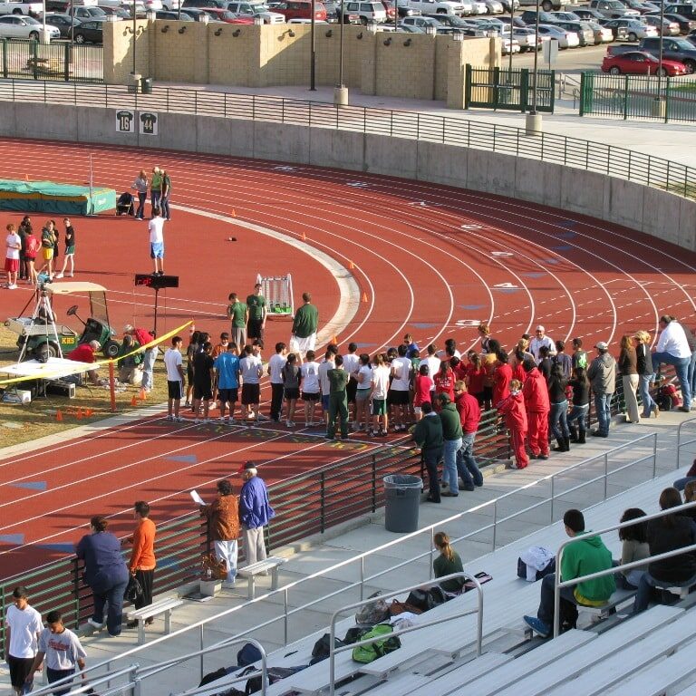 A track and field event is underway at the newly finished Kingsburg High School stadium. The red track is lined with competitors and spectators. In the foreground, people are watching from the new grandstands. A large parking lot is visible in the background.