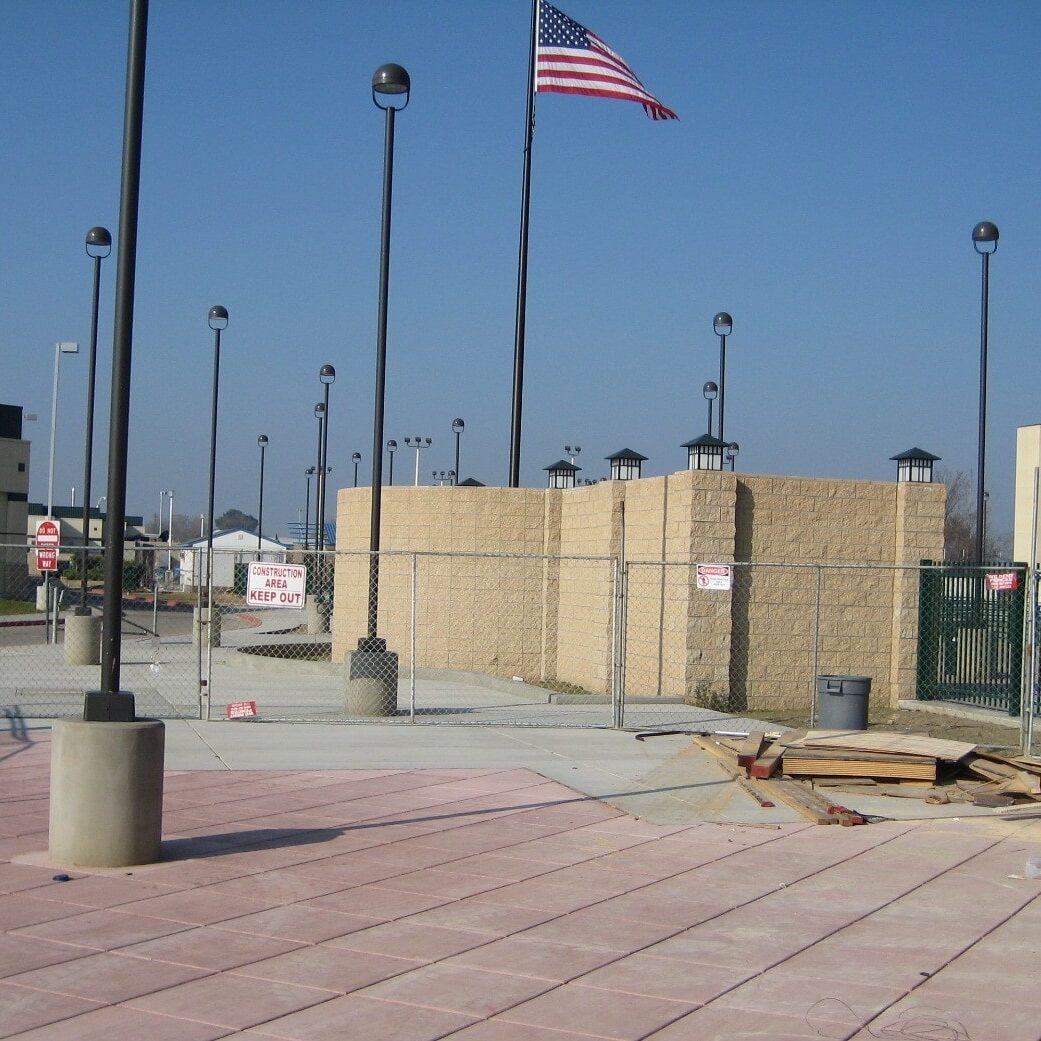 A wide view of the entrance area of the new Kingsburg High School football stadium. A chain-link fence separates the walkway from the construction site. A large American flag flies from a tall flagpole, and a sign on the fence reads "CONSTRUCTION SITE, KEEP OUT".