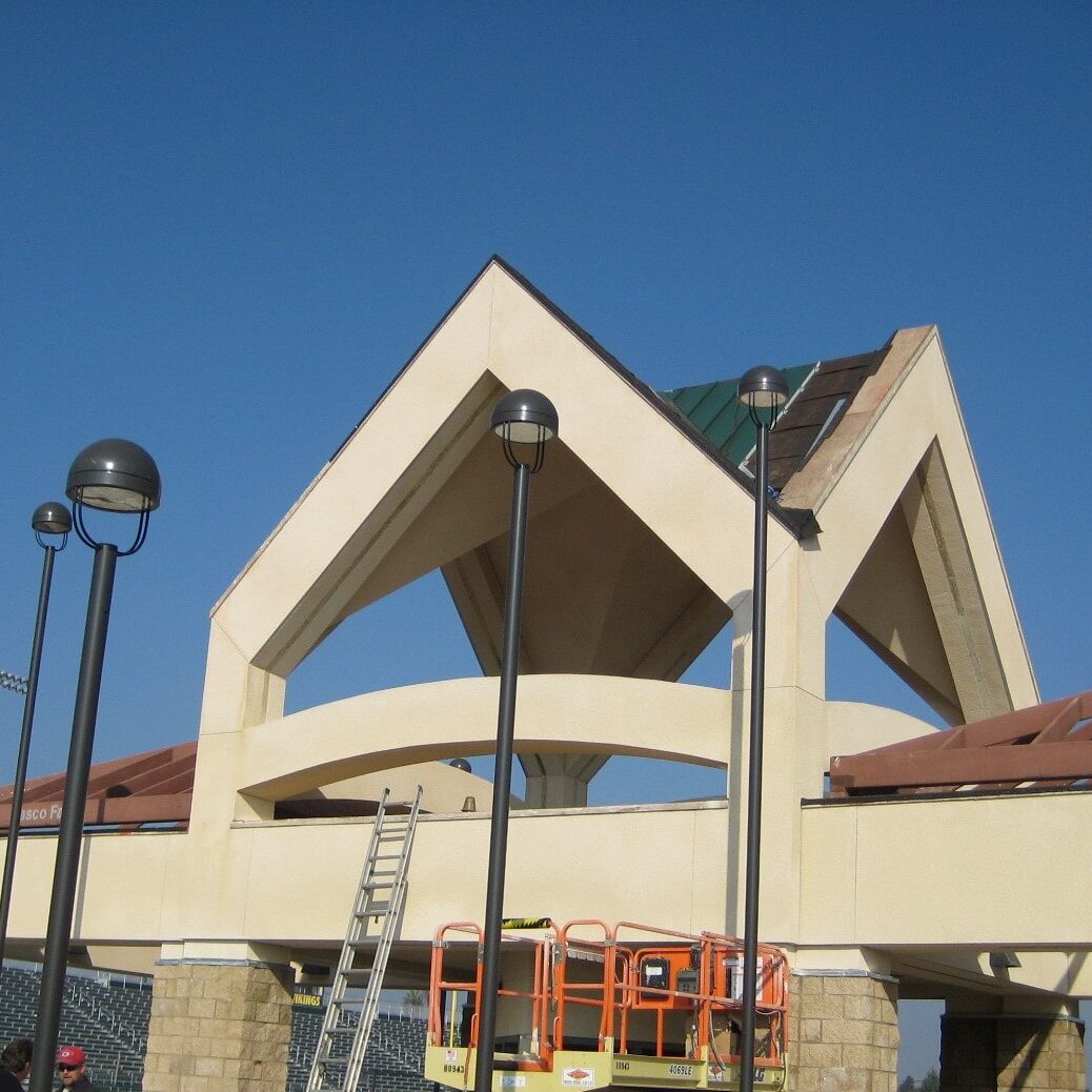 A low-angle shot of a new building at Kingsburg High School's stadium, possibly the concession stand or ticket office. It has a distinctive gabled roofline and is surrounded by black lamp posts. A lift machine is visible next to the building.