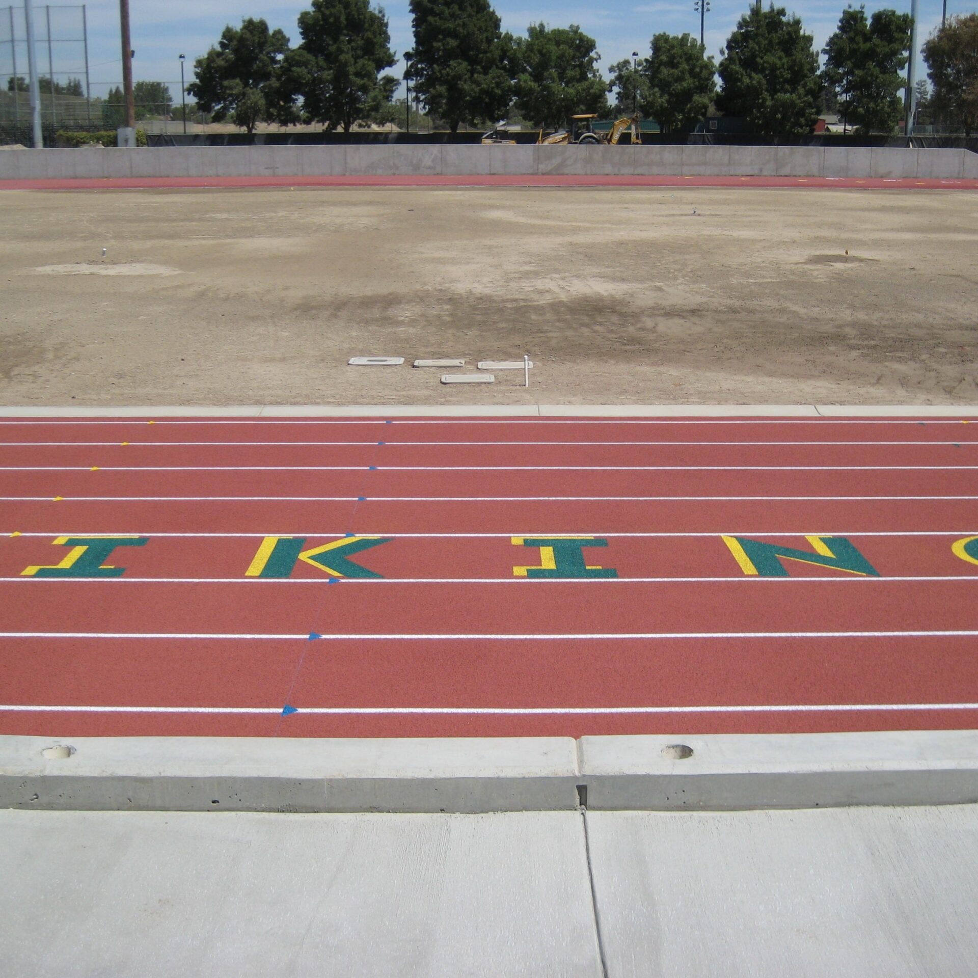 A close-up view of the new red running track at Kingsburg High School's football stadium. The word "VIKINGS" is spelled out in large green and yellow letters on the track. In the background, the ground is dirt and a small tractor is visible.