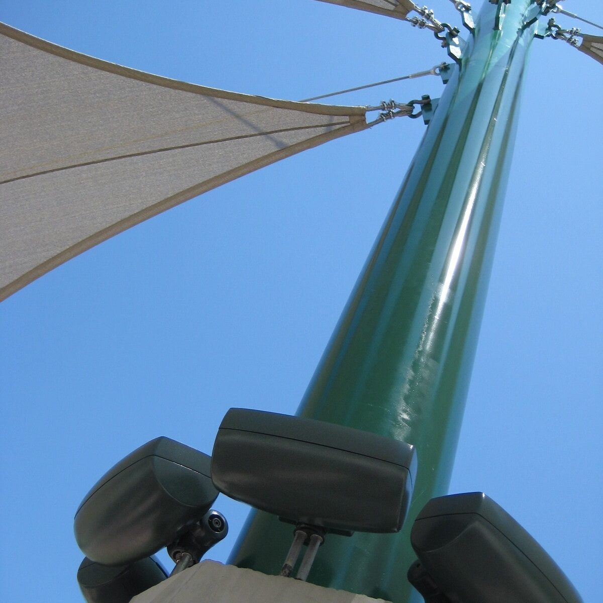 An upward-facing shot of a green pole with several speakers attached to it, possibly at the Kingsburg High School softball stadium. A white fabric canopy is attached to the top of the pole against a clear blue sky.