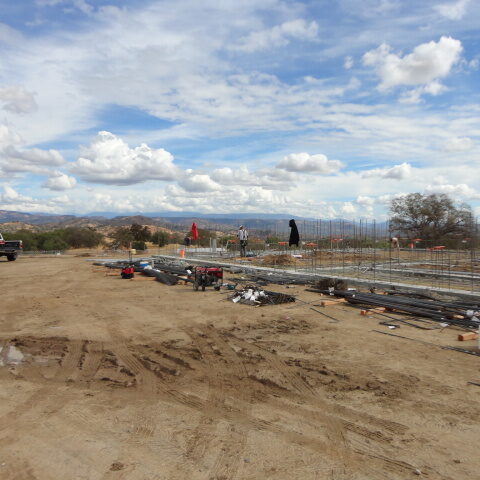 An outdoor construction site with a large, dirt lot in the foreground. In the background, the ground has been leveled and a concrete foundation with rebar extending upwards is visible. Beyond that are rolling hills and a partly cloudy sky. A white truck is parked on the left.