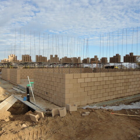An outdoor construction site with partially built block walls. The walls are made of brown bricks and have rebar extending out of the top. Scaffolding is set up behind the walls, and the background shows a row of similar brick structures.