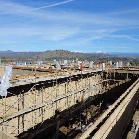 A slightly elevated, wide-angle exterior shot of the construction of a building at Minarets High School. The building's walls are made of light-colored cinder blocks, and the roof is not yet completed. Scaffolding is set up around the structure. In the distance, a scenic landscape with rolling hills and snow-capped mountains under a clear blue sky is visible. A few workers are seen on the roofline. The overall scene captures the building in an unfinished state with a beautiful natural backdrop.
