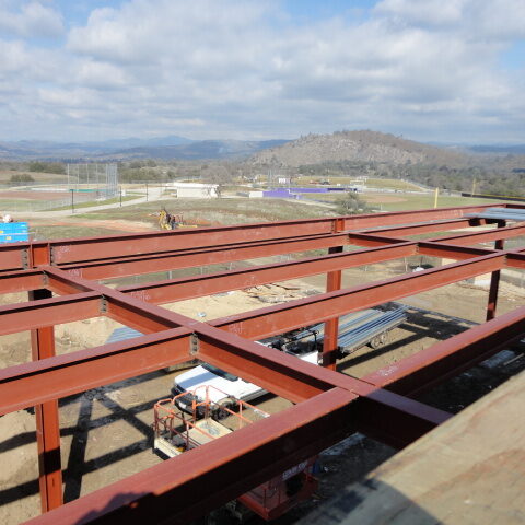 A top-down view of a construction site with the steel frame of a building in the foreground. The red metal beams form a grid pattern. In the distance, there are baseball fields, other buildings, and a hilly, rural landscape.