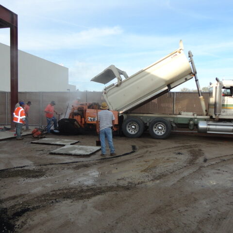 A construction site where a crew is paving. A large dump truck is unloading black asphalt into a paving machine. A few workers in orange vests are standing near the machine, and the ground is covered in fresh asphalt.