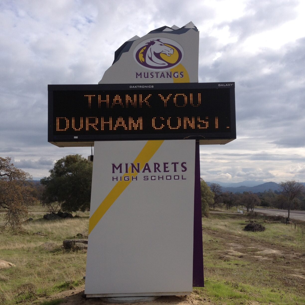 A low-angle, exterior shot of the electronic marquee sign for Minarets High School. The sign is in a grassy field with trees and a road visible behind it. The top of the sign features the school's "Mustangs" mascot logo. The electronic display screen reads "THANK YOU DURHAM CONST.". Below the screen, the sign has the text "MINARETS HIGH SCHOOL" in a modern font with a yellow and purple design. The sky is filled with white and gray clouds. In the distance, hills and mountains are visible.