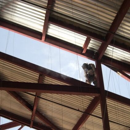 A worker, viewed from below, is welding on the red steel framework of a roof. Sparks are visible, and the roof deck is made of corrugated metal sheets. A clear blue sky is visible through the gaps in the frame.