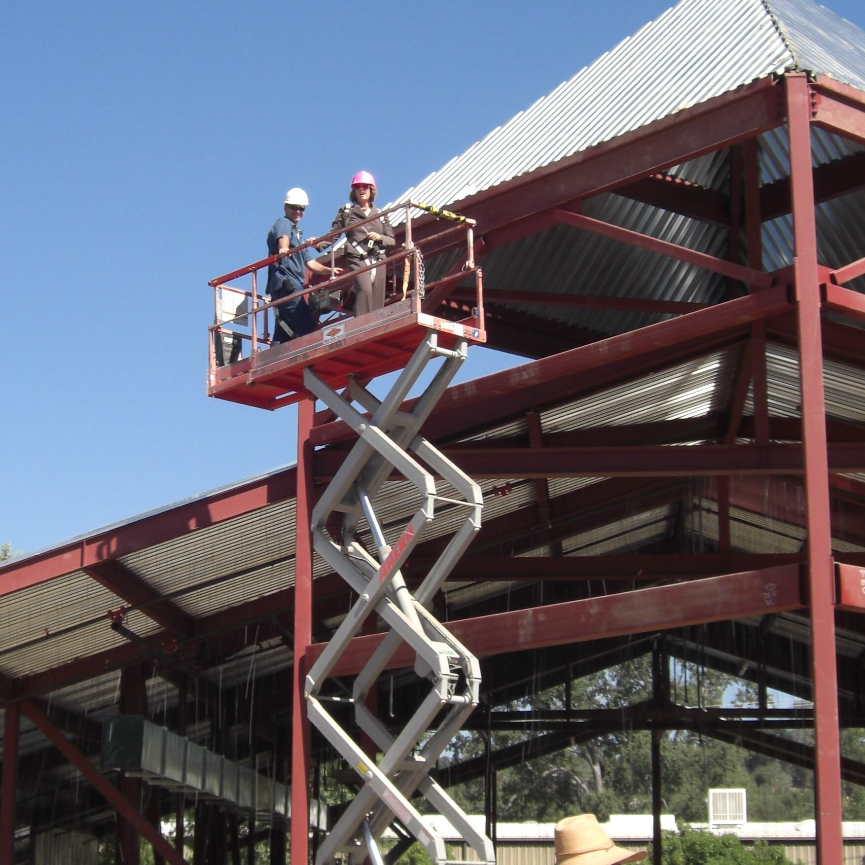 Two people in hard hats, one with a pink hard hat, are standing on the platform of a scissor lift. They are elevated to the level of the red steel building frame. A man in a straw hat is standing below, with his back to the camera.