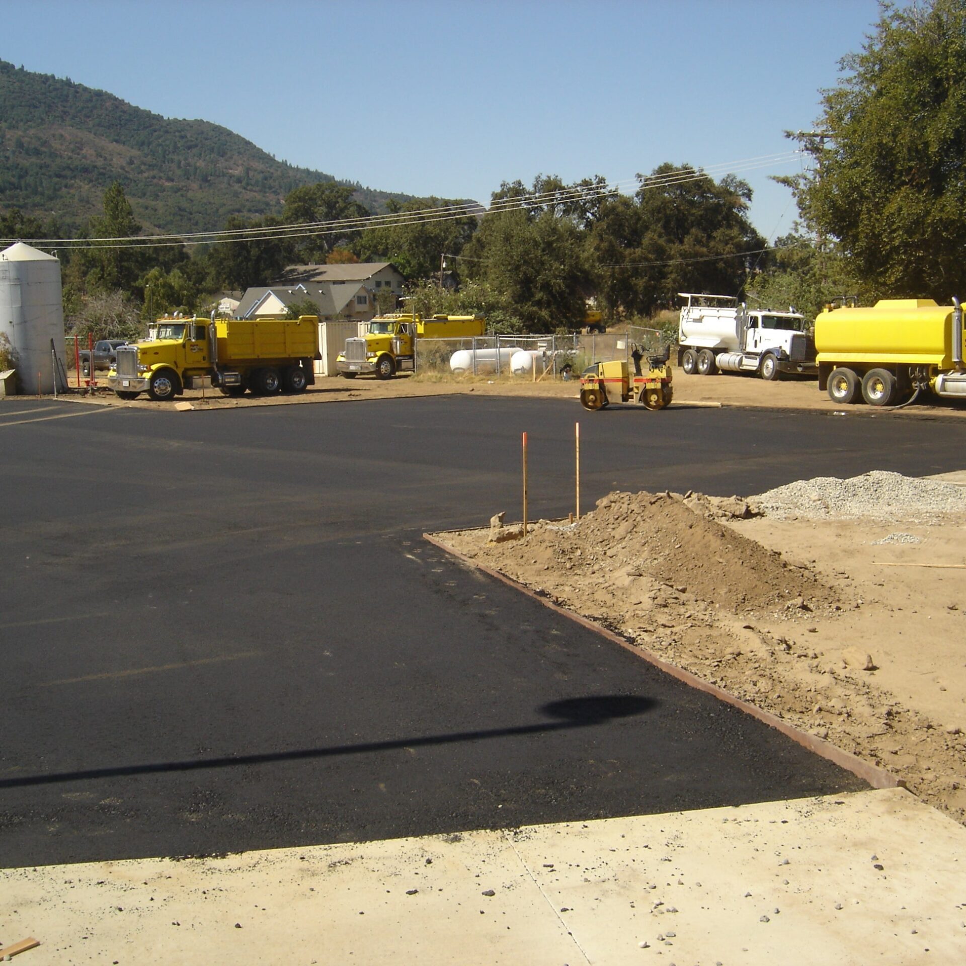 An outdoor scene showing a newly paved asphalt lot, possibly for a parking area. In the background, several yellow construction vehicles, including a dump truck and a roller, are parked. There is a pile of dirt and gravel on the edge of the lot, and a mountain is in the distance.