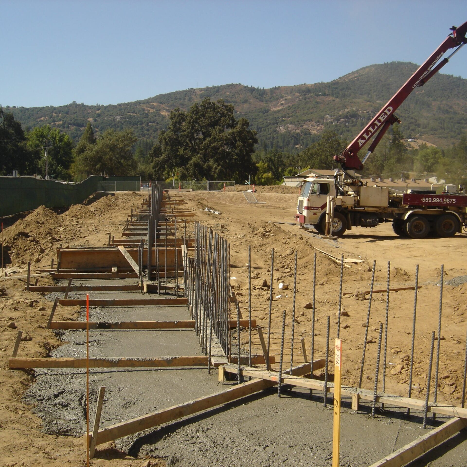 A construction site showing a concrete footing for a masonry wall. The concrete has been poured into wooden forms, and vertical rebar rods extend upwards. A concrete pump truck with a long boom is in the background, set against a hilly landscape.