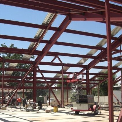 A large red steel frame of a building under construction. The frame is composed of I-beams and trusses. In the background, a person on a scissor lift is working on the structure. The ground is a concrete slab.