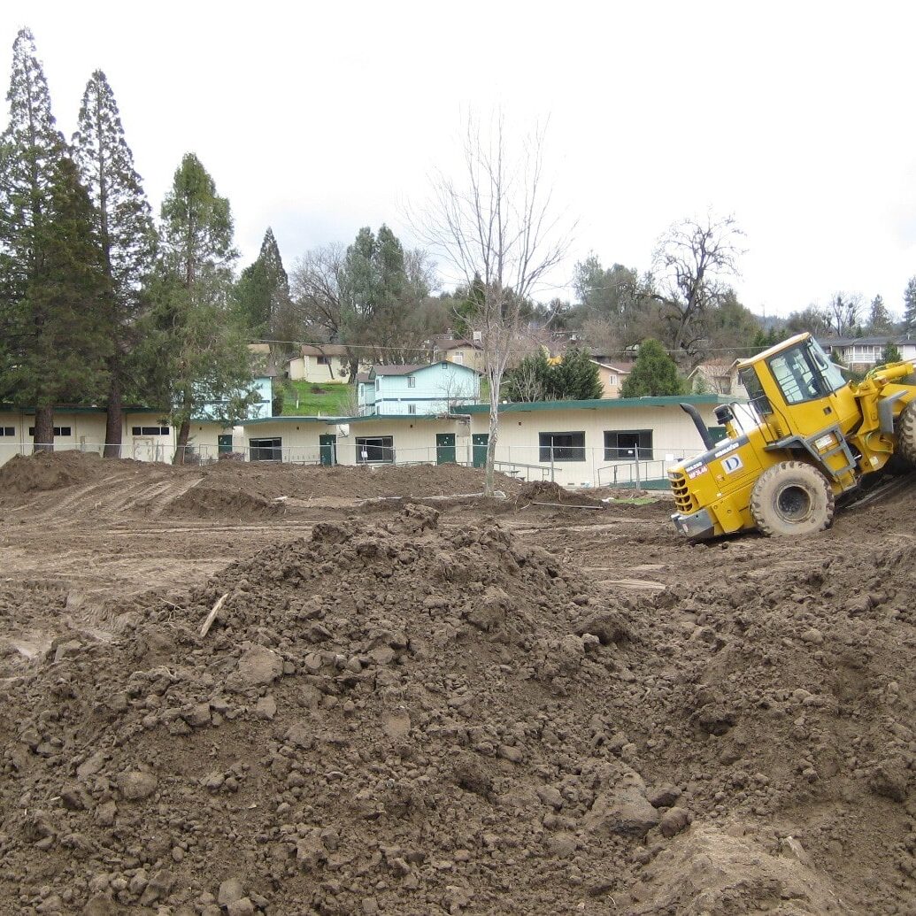 A construction site with large mounds of dirt and a yellow front-end loader parked on a dirt pile. In the background, there is an existing one-story school building with a green roof and a row of trees.