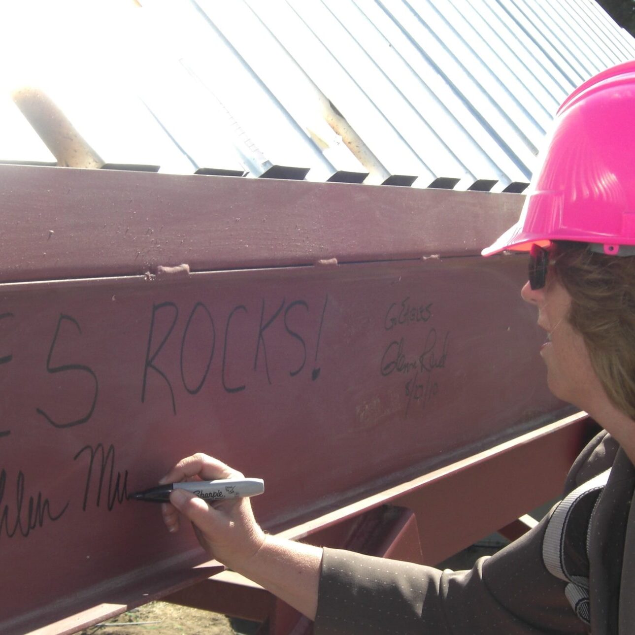 A person in a pink hard hat is signing their name with a black marker on a red metal I-beam. Other signatures are also visible on the beam. The background shows a metal roof structure.
