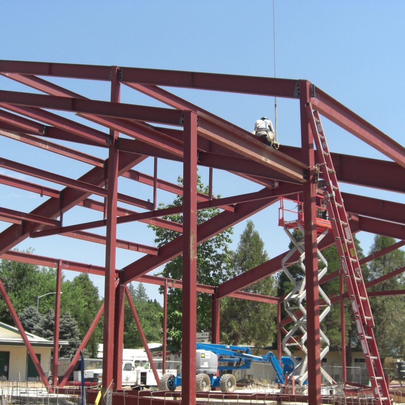 A red steel frame of a building is being erected against a clear blue sky. A worker is high on the structure, and a ladder and a blue scissor lift are positioned below.