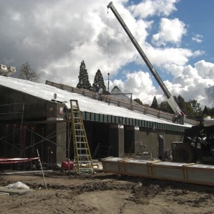 The exterior of a building under construction. The walls are covered in a gray material and the roof has a new metal deck. A crane is positioned to lift materials onto the roof, and a large flatbed truck is parked with more roofing materials.